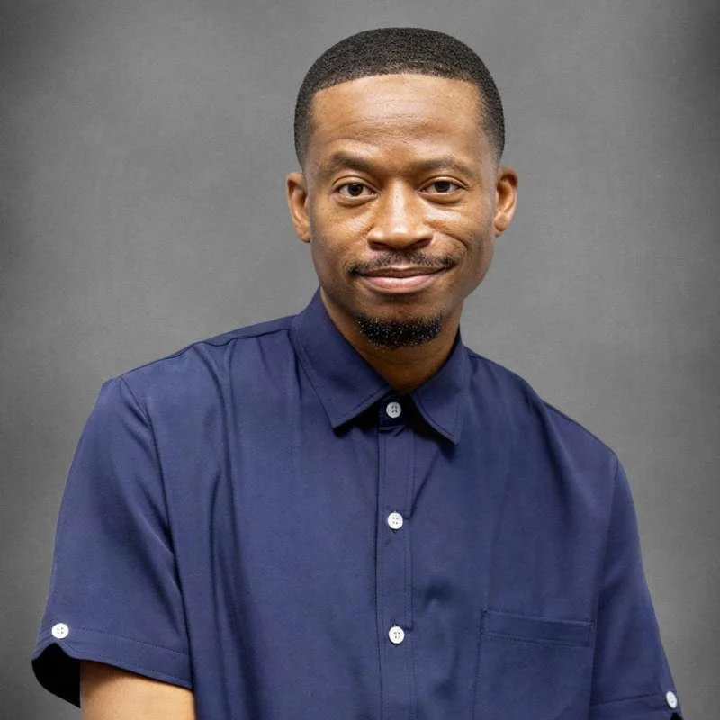 Portrait of a man wearing a dark blue short-sleeved shirt, standing in front of an abstract painting with vertical black, white, and blue brushstrokes.