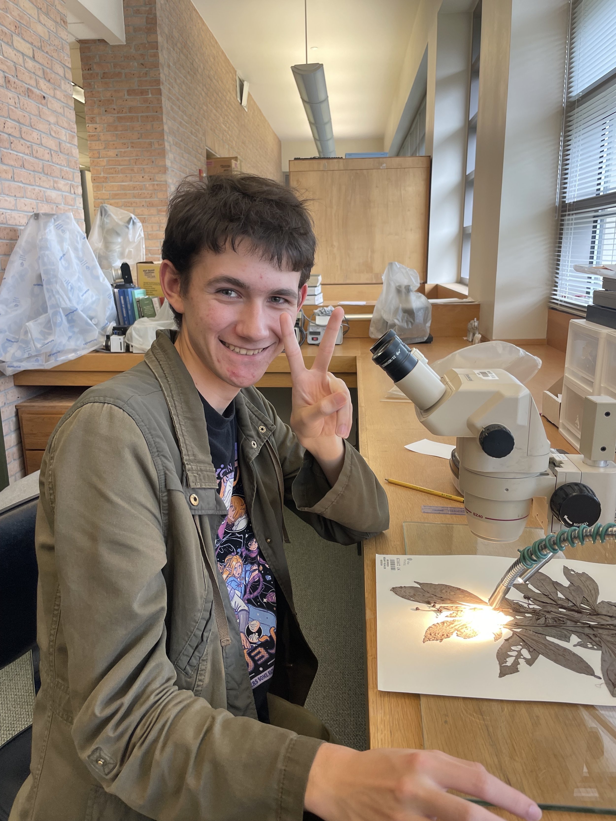 Nolan gives a peace sign while looking at an herbarium specimen under a dissecting scope