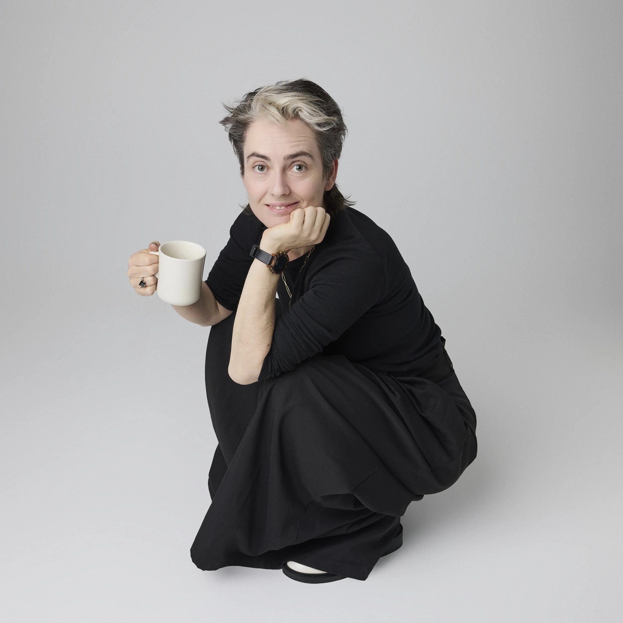 Person with short gray hair wearing black clothing, squatting on the floor, holding a white mug, and looking at the camera.