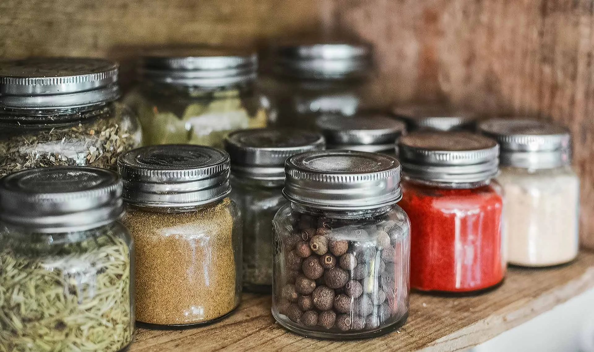 Assorted spices and herbs in glass jars with metal lids on a wooden shelf.