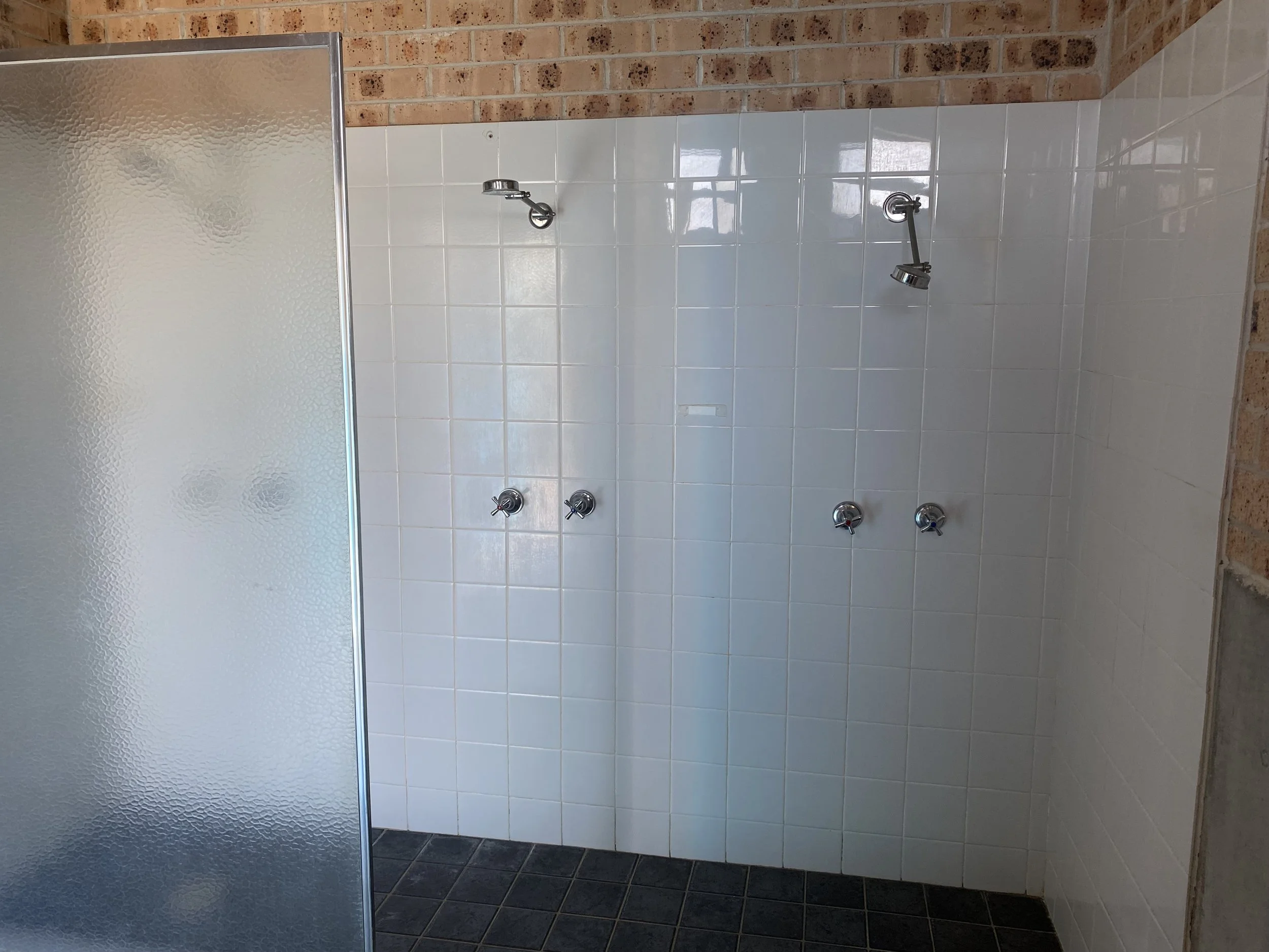 Public shower area with multiple showerheads and a partially frosted glass shower partition on the left, with white tiled wall and brick wall in the background.