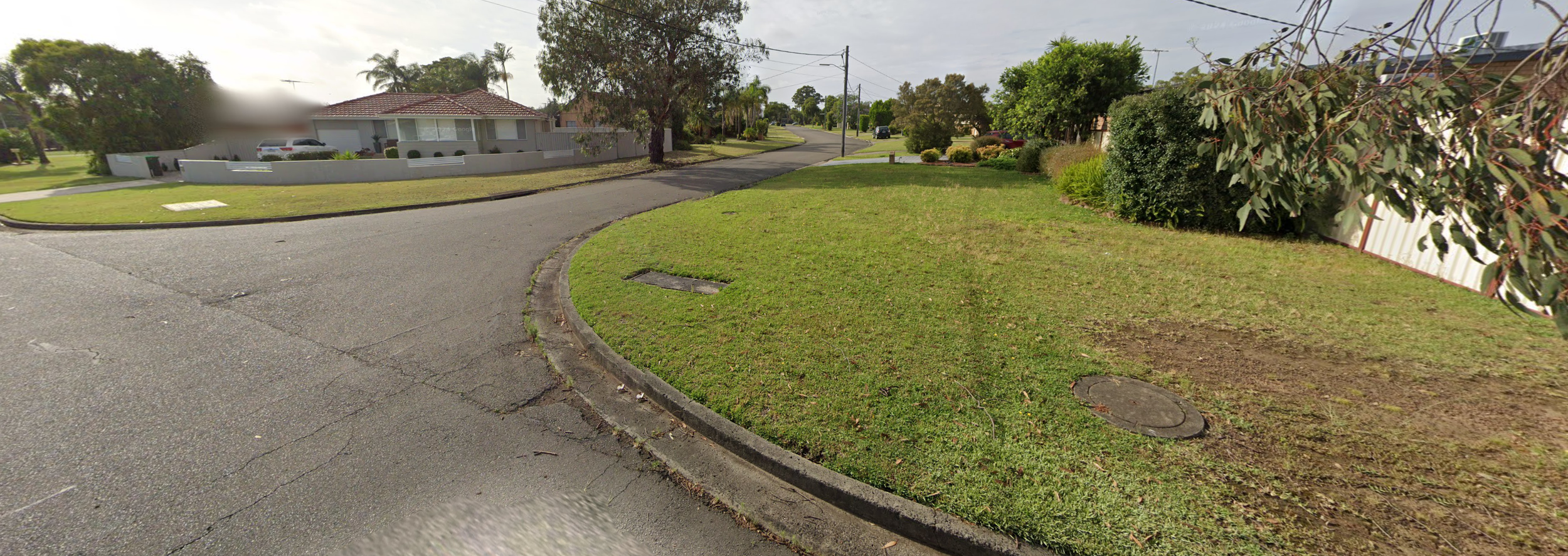 Suburban neighborhood street with grassy lawns, trees, and houses, showing a curved road and a cloudy sky.