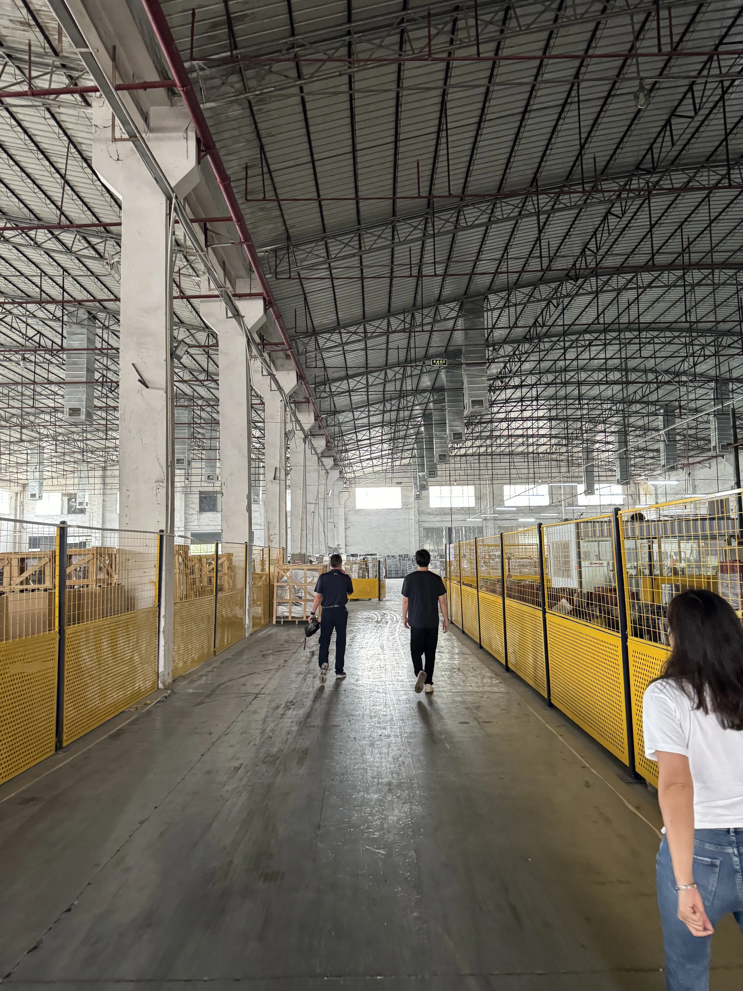 Three people walk through an industrial warehouse with a high ceiling, concrete floor, and yellow safety fences along the sides.