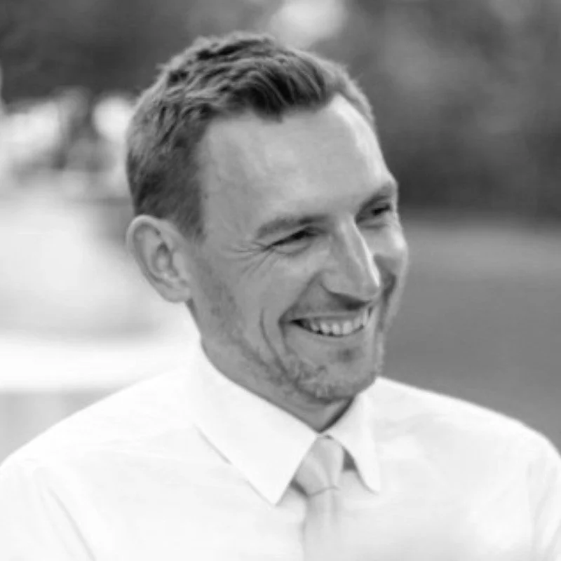 Black and white photo of a smiling man in a collared shirt and tie, outdoors.