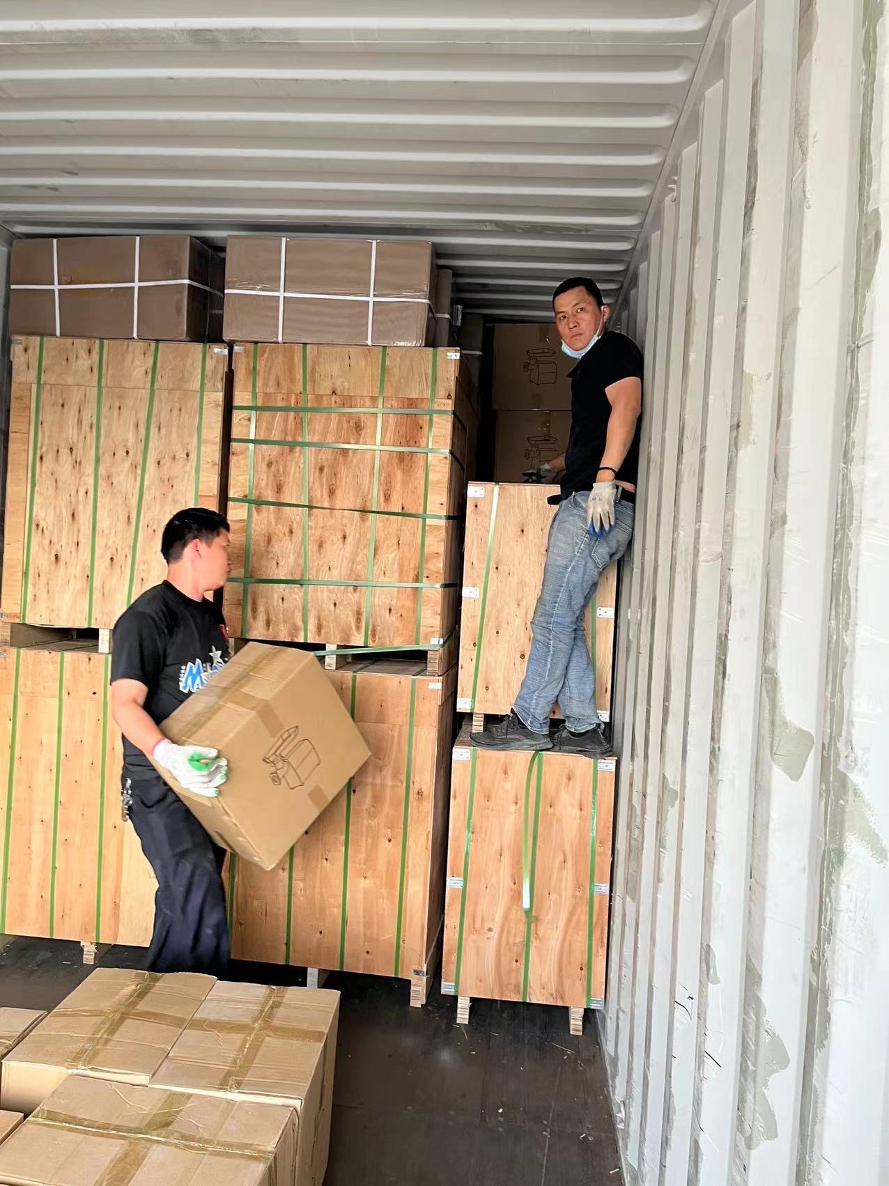 Two men are loading or unloading cardboard boxes and wooden crates inside a shipping container. One man is carrying a box, and the other is standing on a crate.