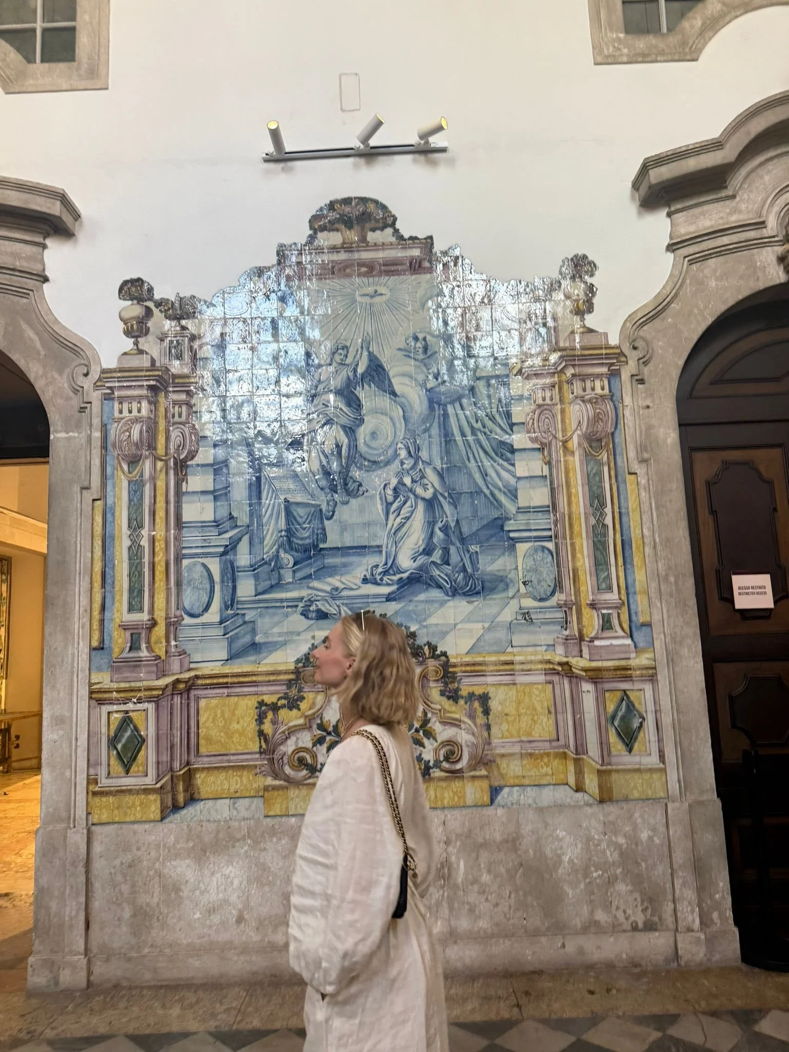 A woman with blonde, wavy hair and a beige coat with a chain strap purse standing in front of a decorative tiled mural in an indoor setting.
