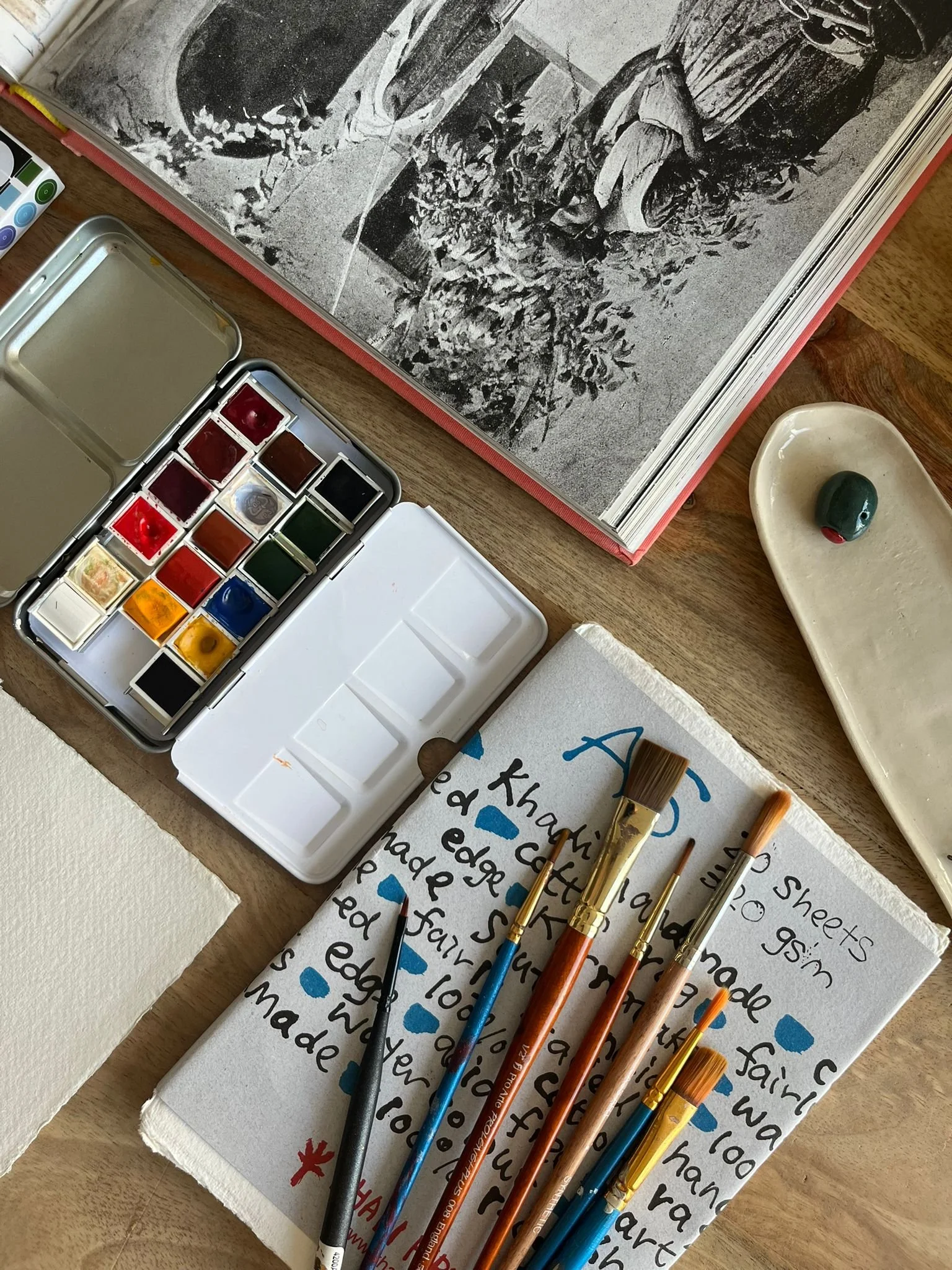 Watercolor paints, paintbrushes, and a notebook with handwritten text and blue paint drops on a wooden table, alongside an open book with black-and-white photographs.