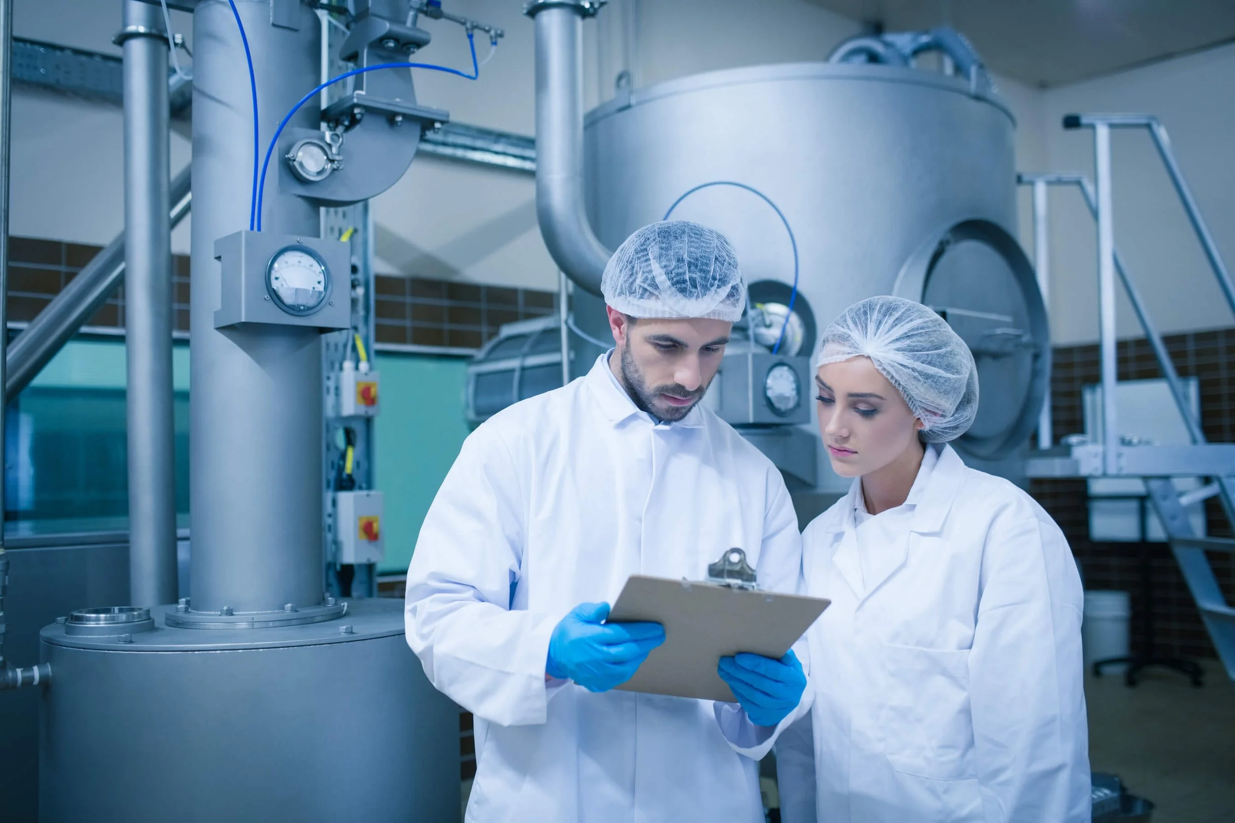 Two people in a laboratory or industrial setting with white coats, hairnets and blue gloves. They are standing next to metallic equipment with tubes and measuring instruments, and one of them is holding a clipboard