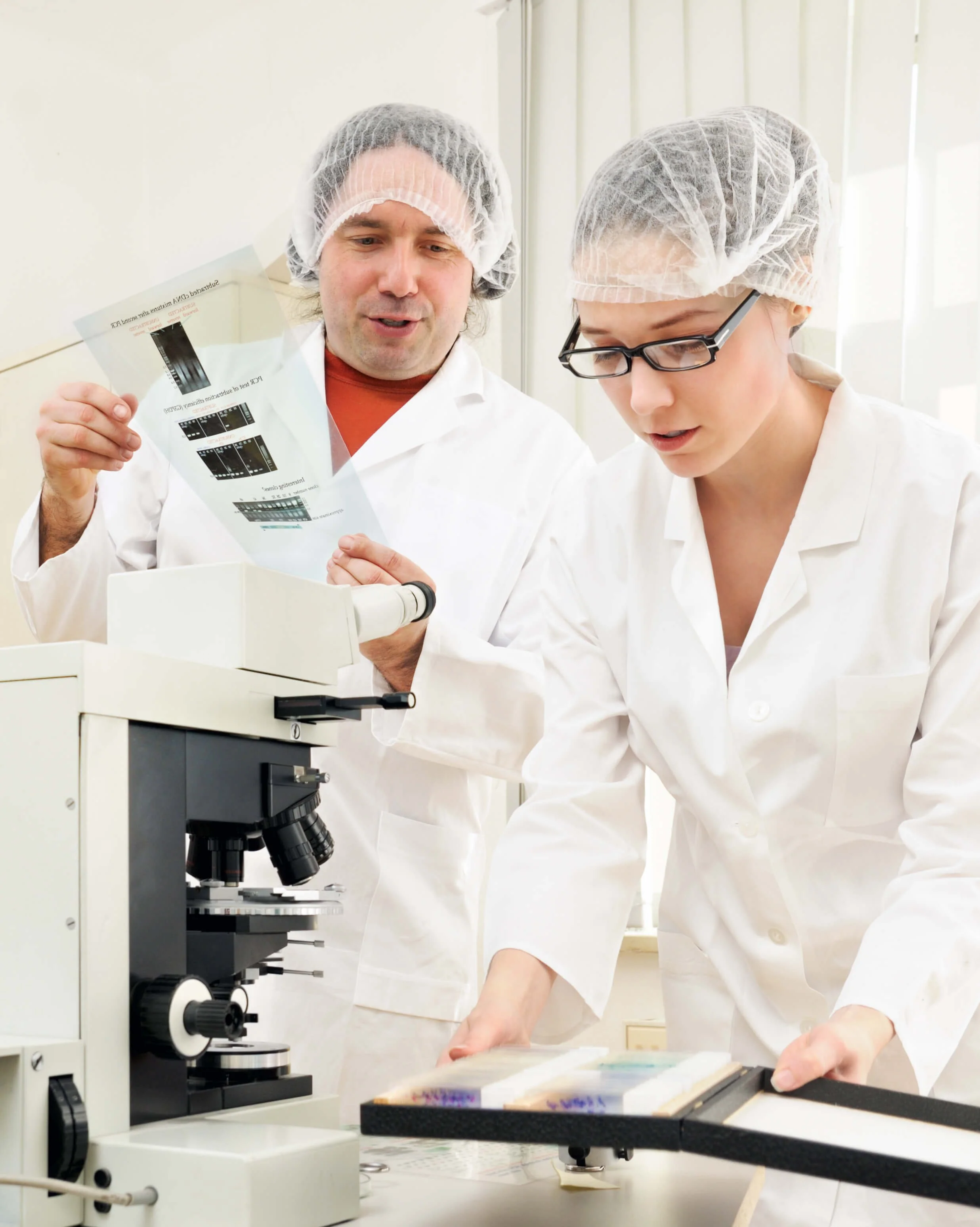 Two people in lab coats with hairnets, one holding a microscope