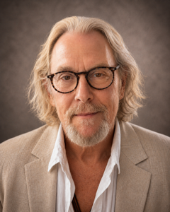 A middle-aged man with gray hair and a beard, wearing a black jacket over a white shirt, looking directly at the camera with a neutral expression against a dark background.