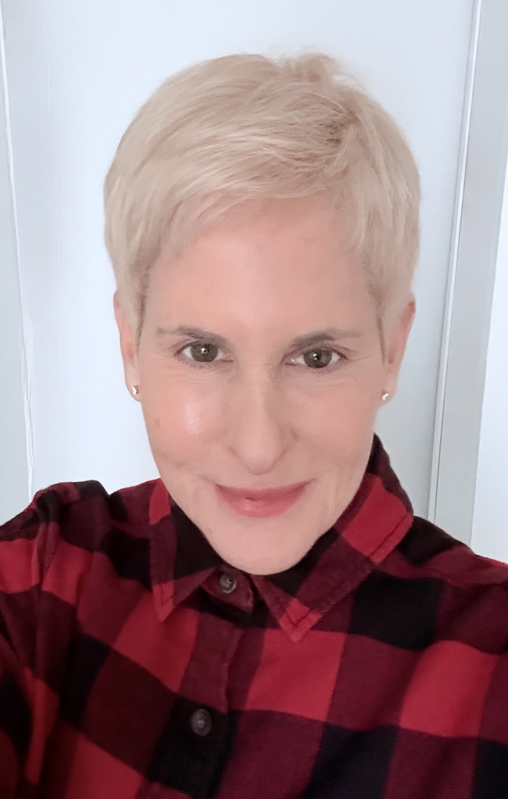 A woman with short dark hair smiling, wearing a beige knit turtleneck sweater, standing indoors near a window with blurred greenery outside.