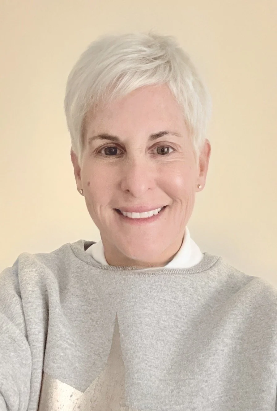 A woman with short dark hair smiling, wearing a beige knit turtleneck sweater, standing indoors near a window with blurred greenery outside.