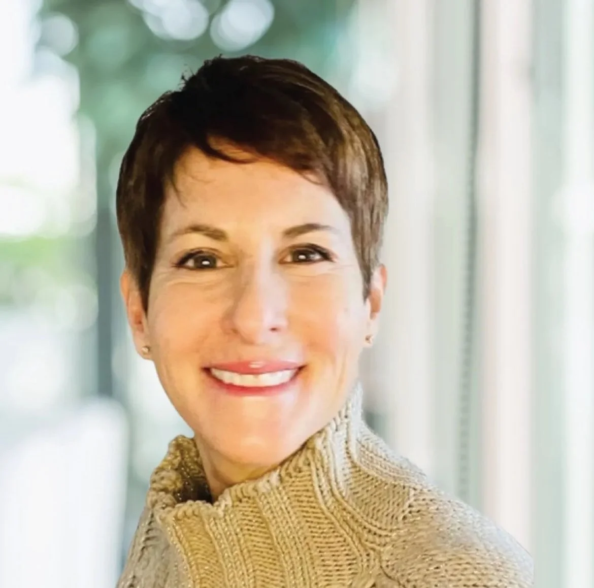 A woman with short dark hair smiling, wearing a beige knit turtleneck sweater, standing indoors near a window with blurred greenery outside.