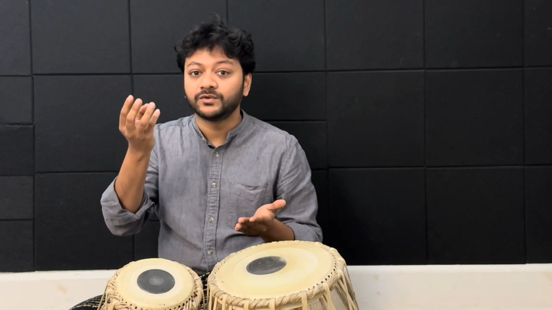 Mir Naqibul Islam playing tablas with a backdrop of black soundproof panels.