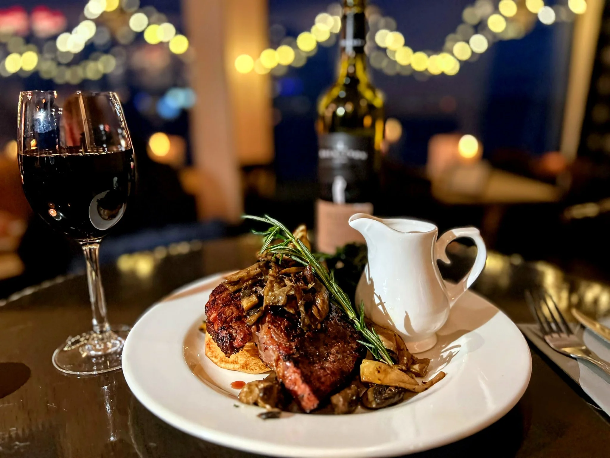 A glass of red wine and a plate with a cooked steak topped with caramelized onions and herbs, accompanied by a small white pitcher, on a dark table with blurred warm decorative lights in the background.