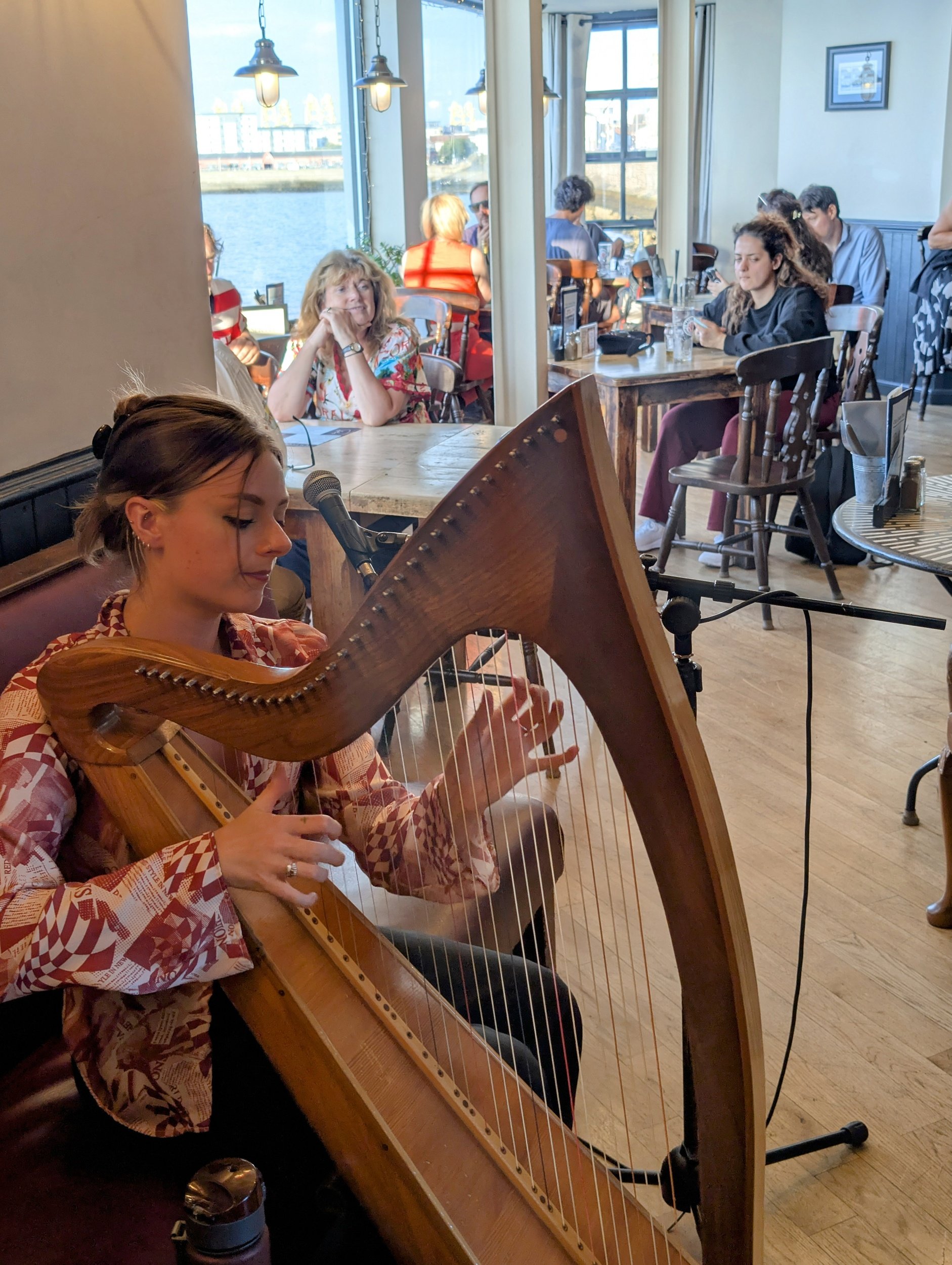 Woman playing harp in a cozy coffee shop with large windows showing water and buildings outside.