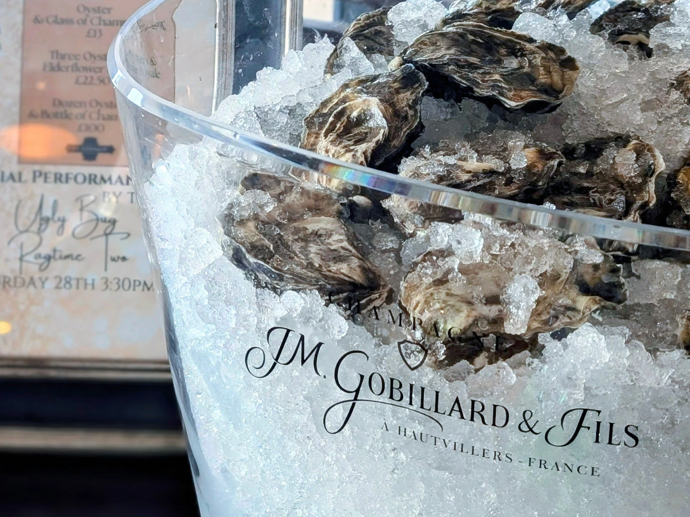 Close-up of a glass filled with oysters on a bed of crushed ice. The glass has decorative black script writing on it and is placed near a menu or informational card in the background.