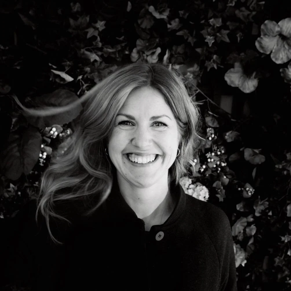 Et svart-hvitt bilde av en smilende kvinne med langt, bølgete hår, iført en mørk jakke, med blomster og blader i bakgrunnen.