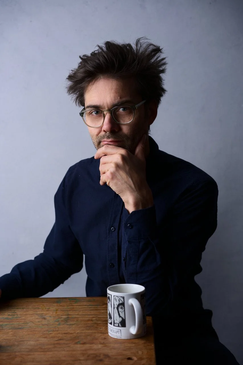 En mann med brunt, rotete hår, iført briller og mørk skjorte sitter ved et bord med en kopp. Han har en tenksom uttrykk og hviler hånden mot haken.