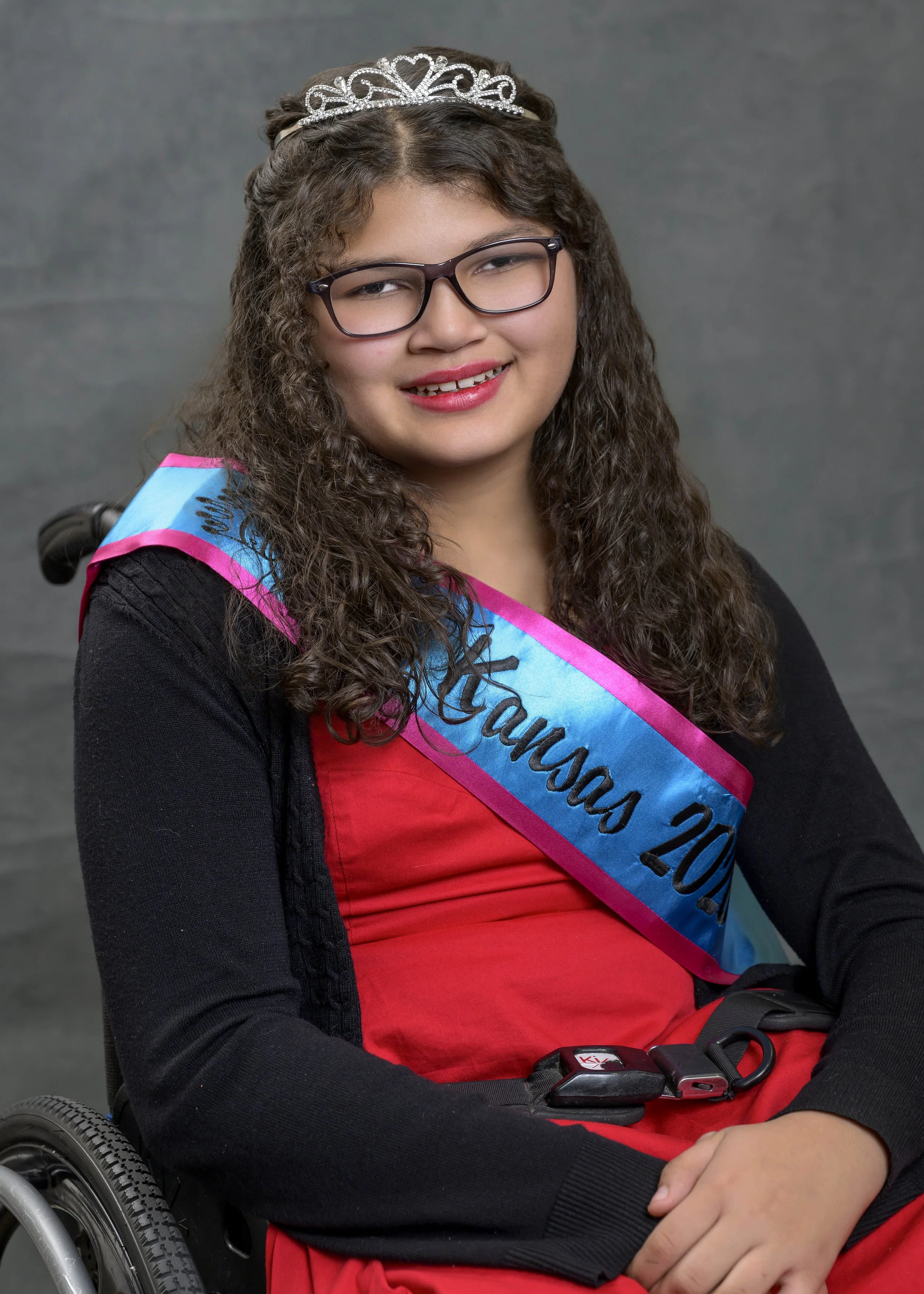 Naomi smiling for her official portrait. She is wearing a teal and pink sash, crown, red dress with black sweater, and black glasses. Naomi is using a manual wheelchair and has long curly brown hair.