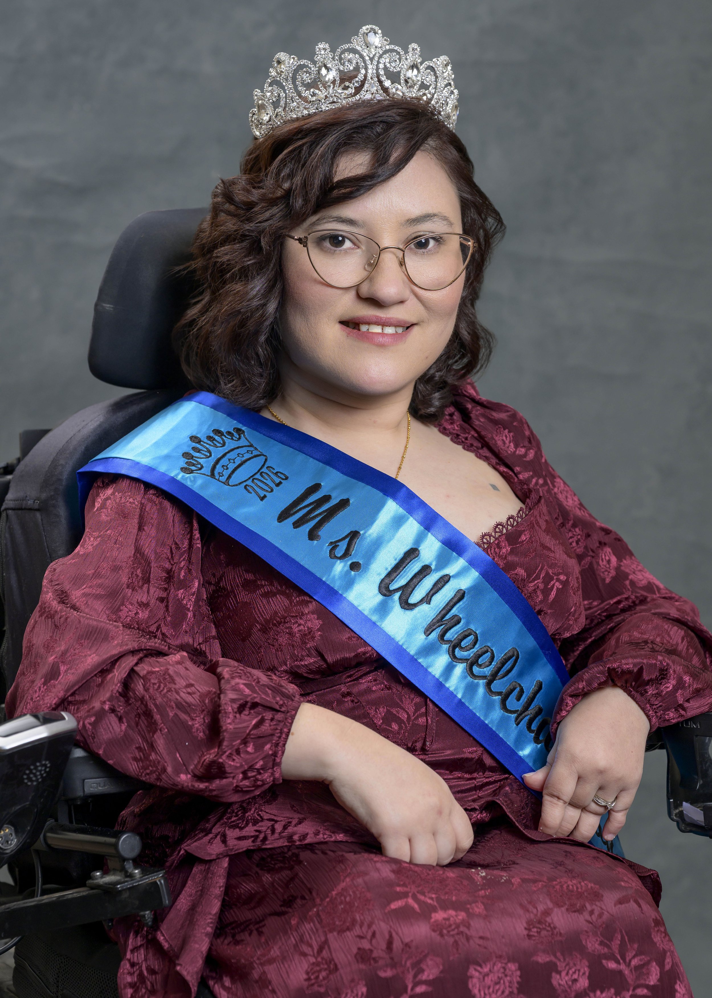 Magda smiling for her portrait. She is wearing her teal and blue sash, crown, burgundy dress, and glasses. Magda is using a black power chair and has shoulder length curly brown hair.