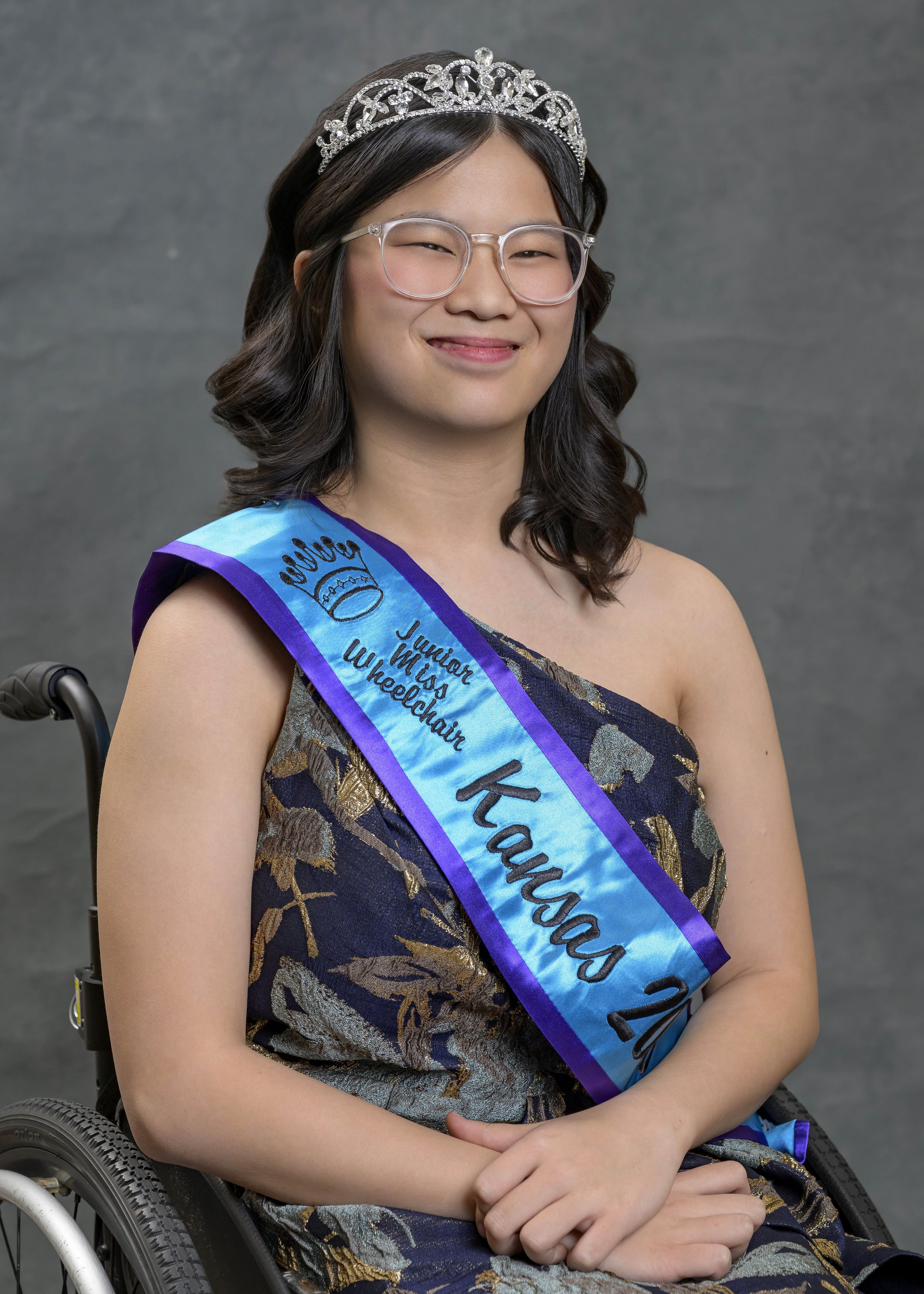 AJ smiling for her portrait. She is wearing a dark blue and gold dress, teal and purple sash, crown, and glasses. AJ is using a manual chair and has wavy long dark brown hair.