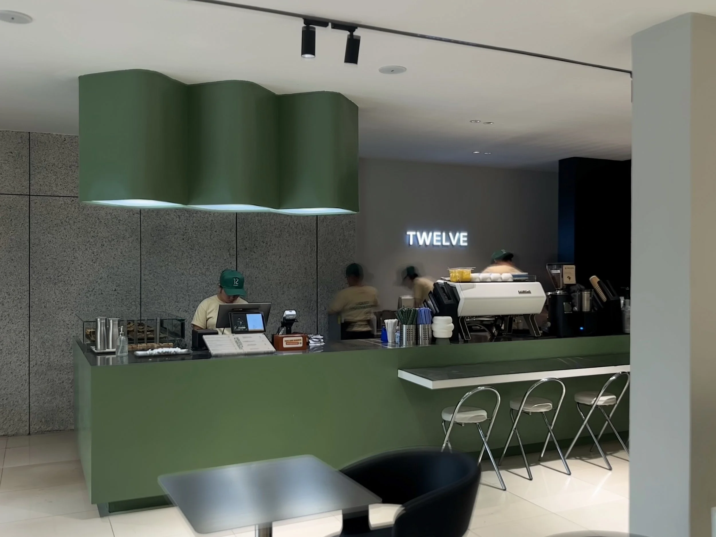 A modern sage green coffee counter at Twelve Coffee Roastery featuring a sculpted wavy green overhead light, baristas at POS and espresso machines, and metallic bar stools.