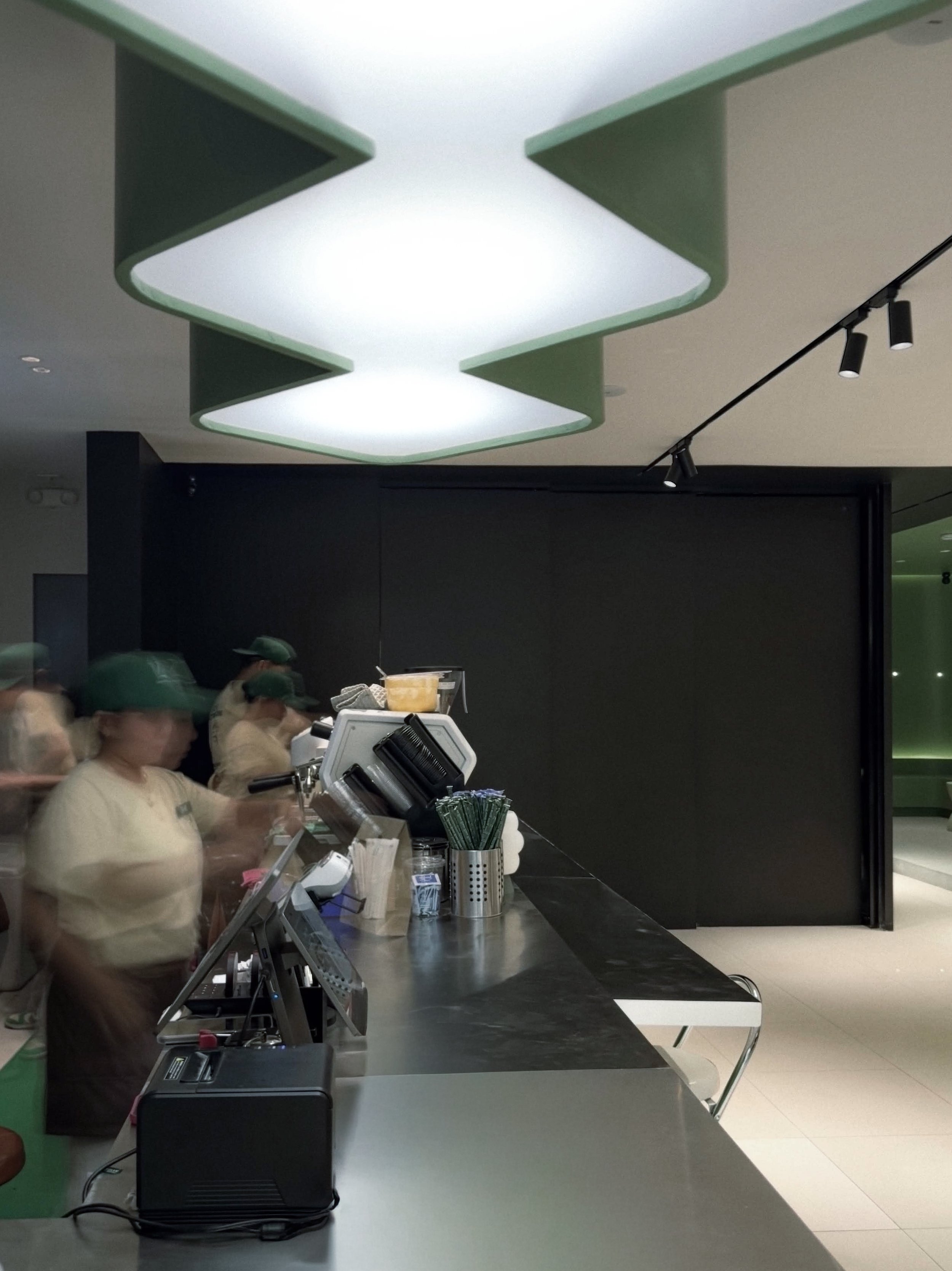 Interior view of Twelve Coffee Roastery showing baristas working behind a stainless steel service counter beneath a unique wavy green ceiling light fixture.