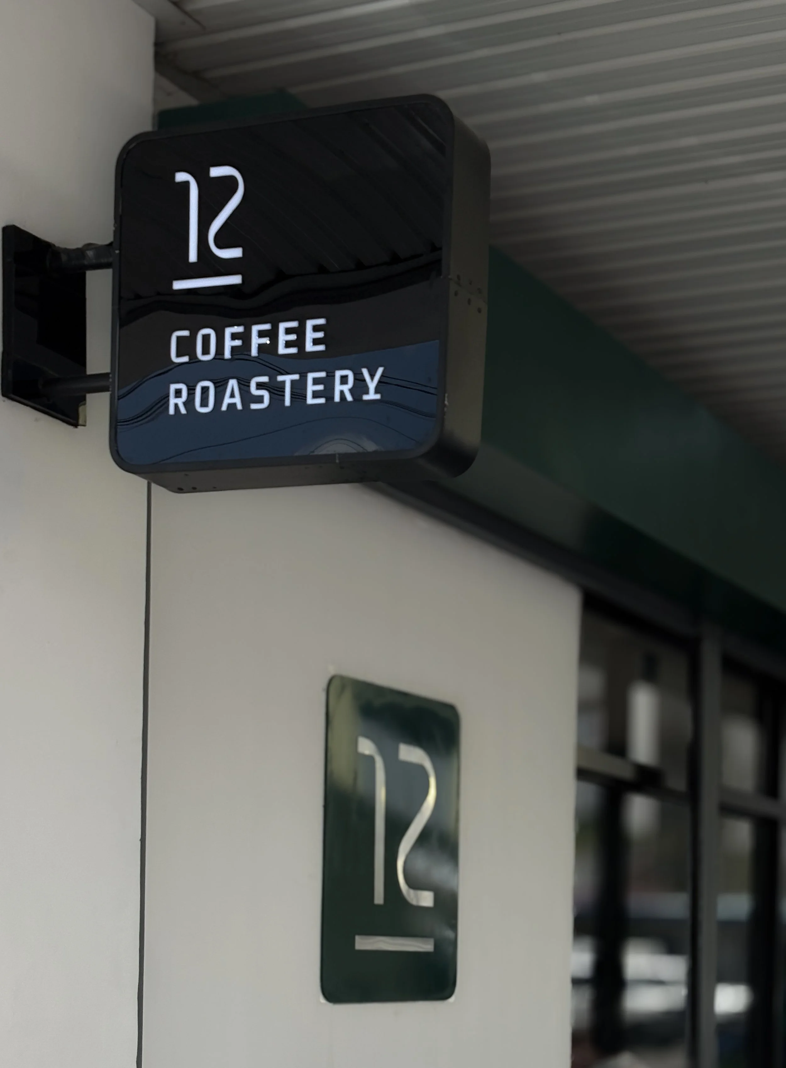Close-up of the exterior branding for Twelve Coffee Roastery, featuring a black illuminated blade sign and a wall plaque with the "12" logo.
