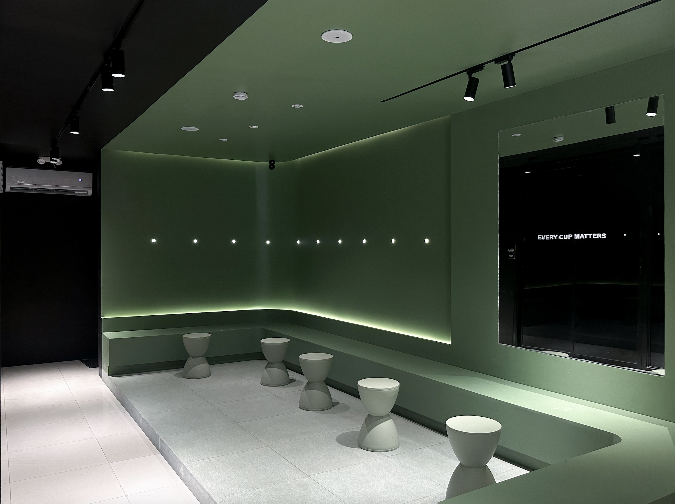 Minimalist green seating nook at Twelve Coffee Roastery featuring built-in benching, light grey hourglass stools, ambient wall lighting, and a window with text reading "EVERY CUP MATTERS."