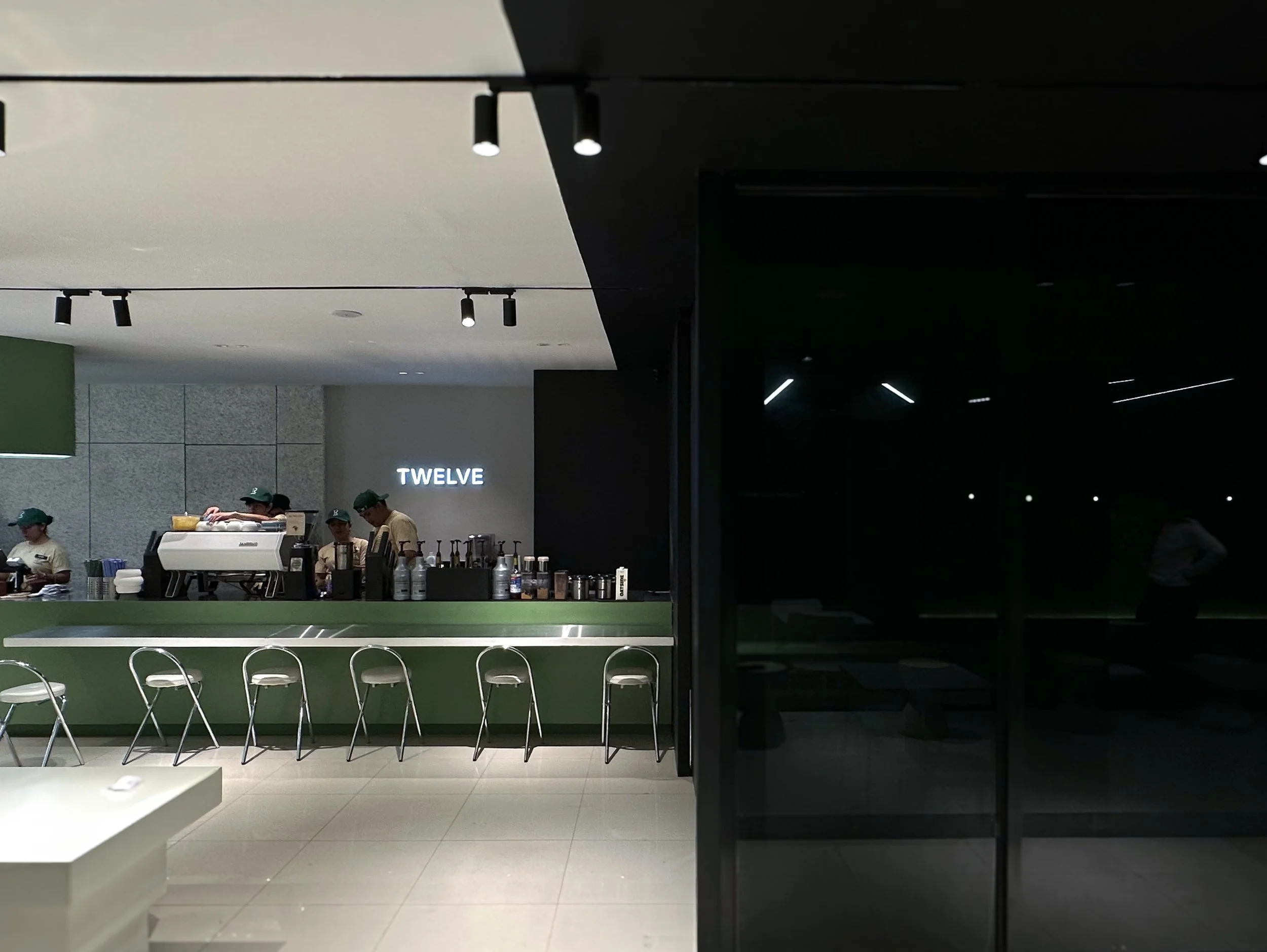 Wide shot of Twelve Coffee Roastery's main espresso bar featuring a sage green counter, metallic bar stools, illuminated "TWELVE" wall logo, and dark architectural accents.
