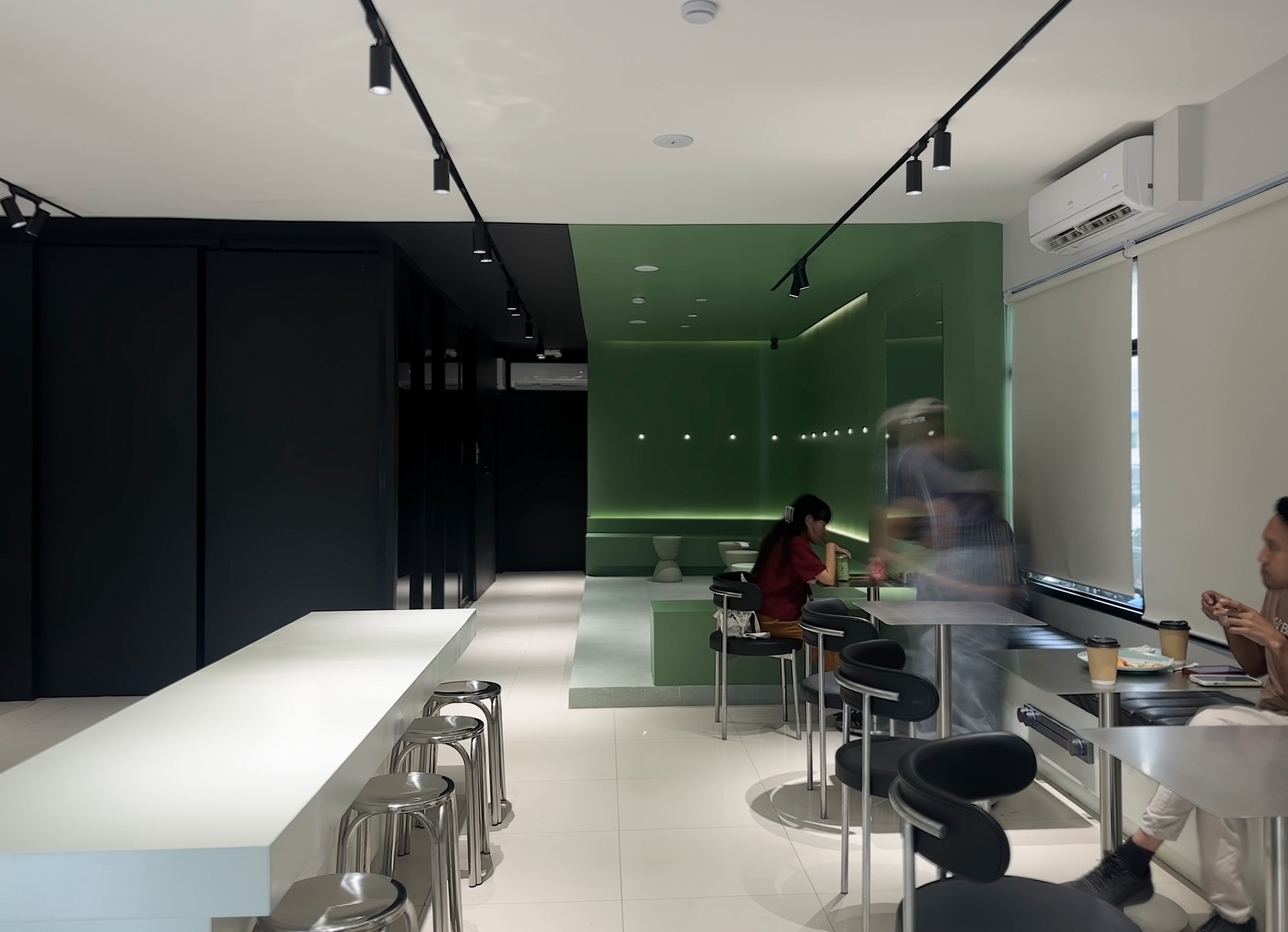 Modern, minimalist coffee shop interior with black, white, and muted green, sage green, color blocking, track lighting, metallic bar stools, and patrons seated at small tables.