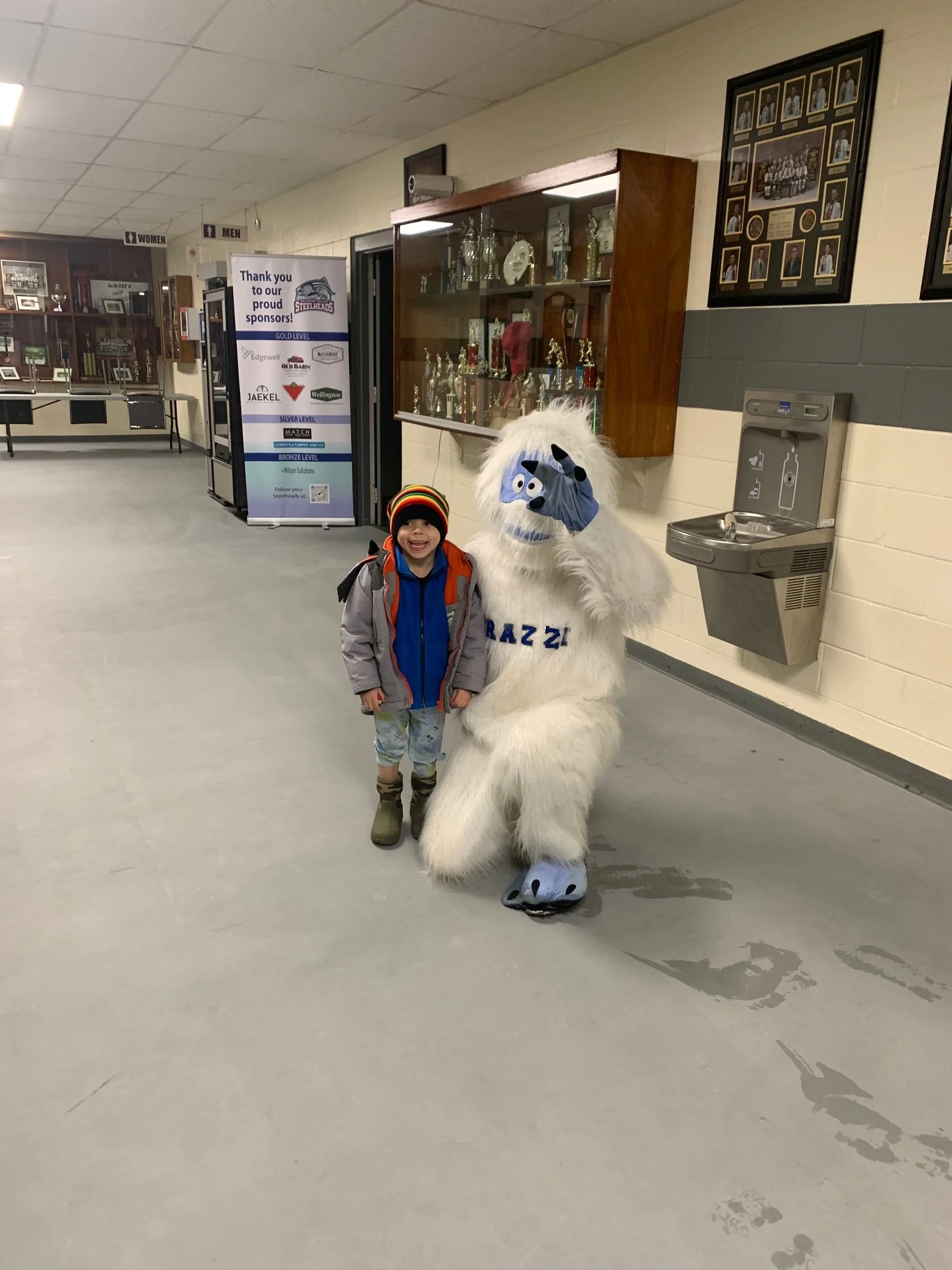 A young boy standing next to a person in a Sasquatch costume, both smiling in an indoor school hallway with trophies and framed photos on the wall.