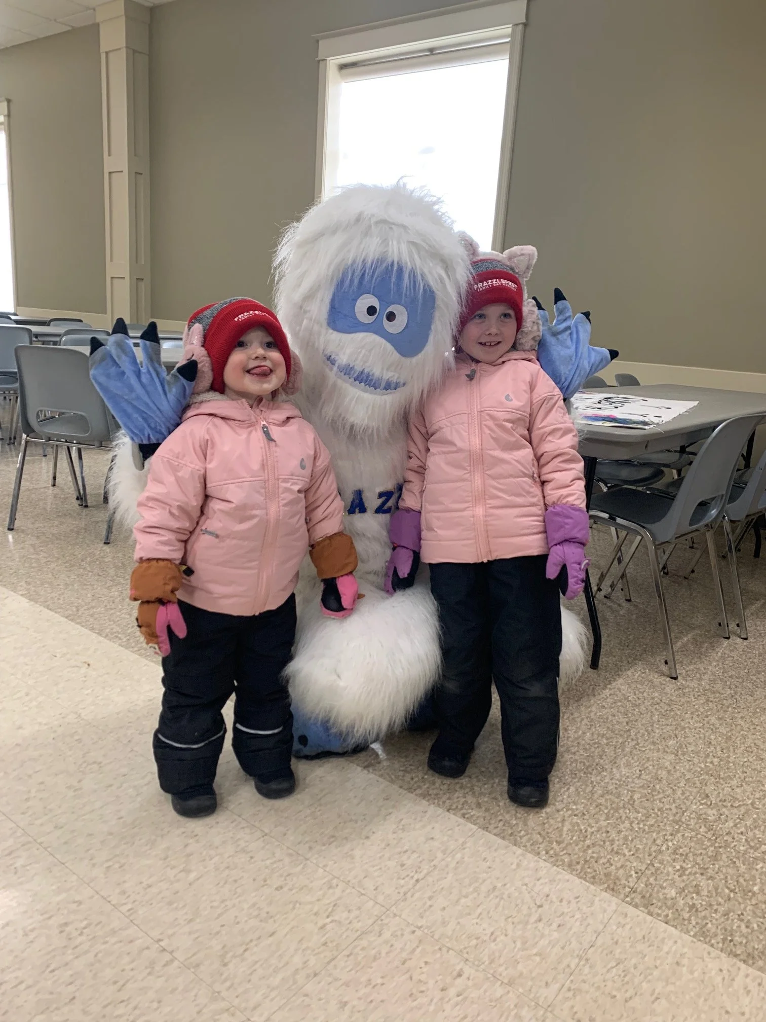 Two young children in pink jackets and winter hats pose with a large blue furry mascot with white hair and beard, and big eyes, inside a room with tables and chairs.