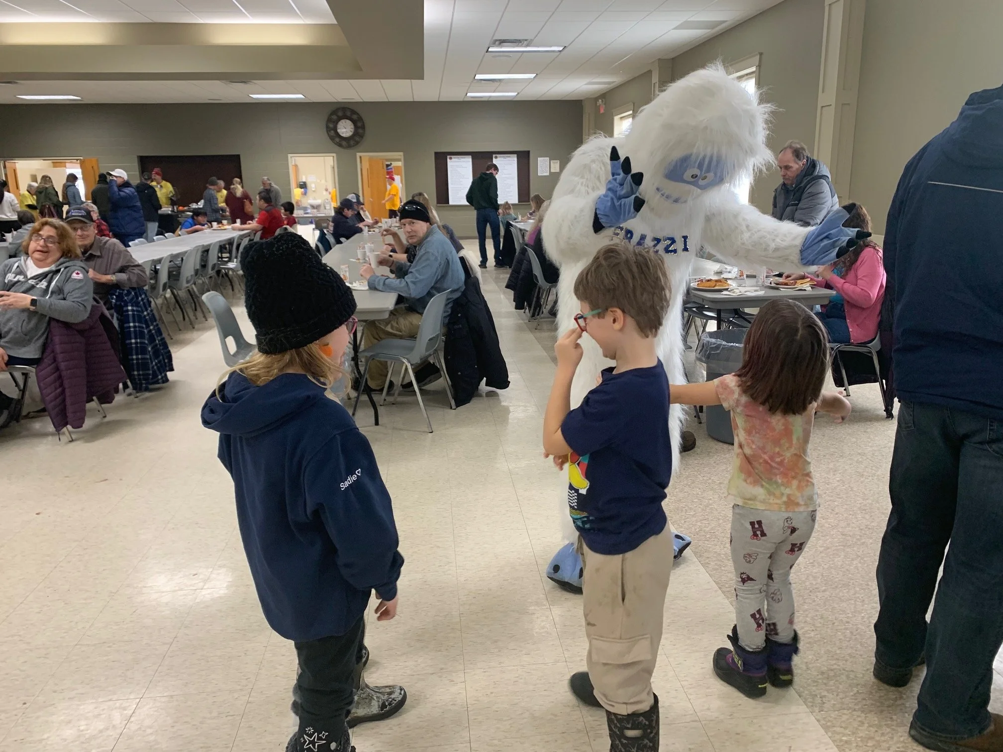 Children interact with a person in a large furry mascot costume resembling a police officer, in a crowded indoor event space with tables, people eating, and a mascot making gestures.