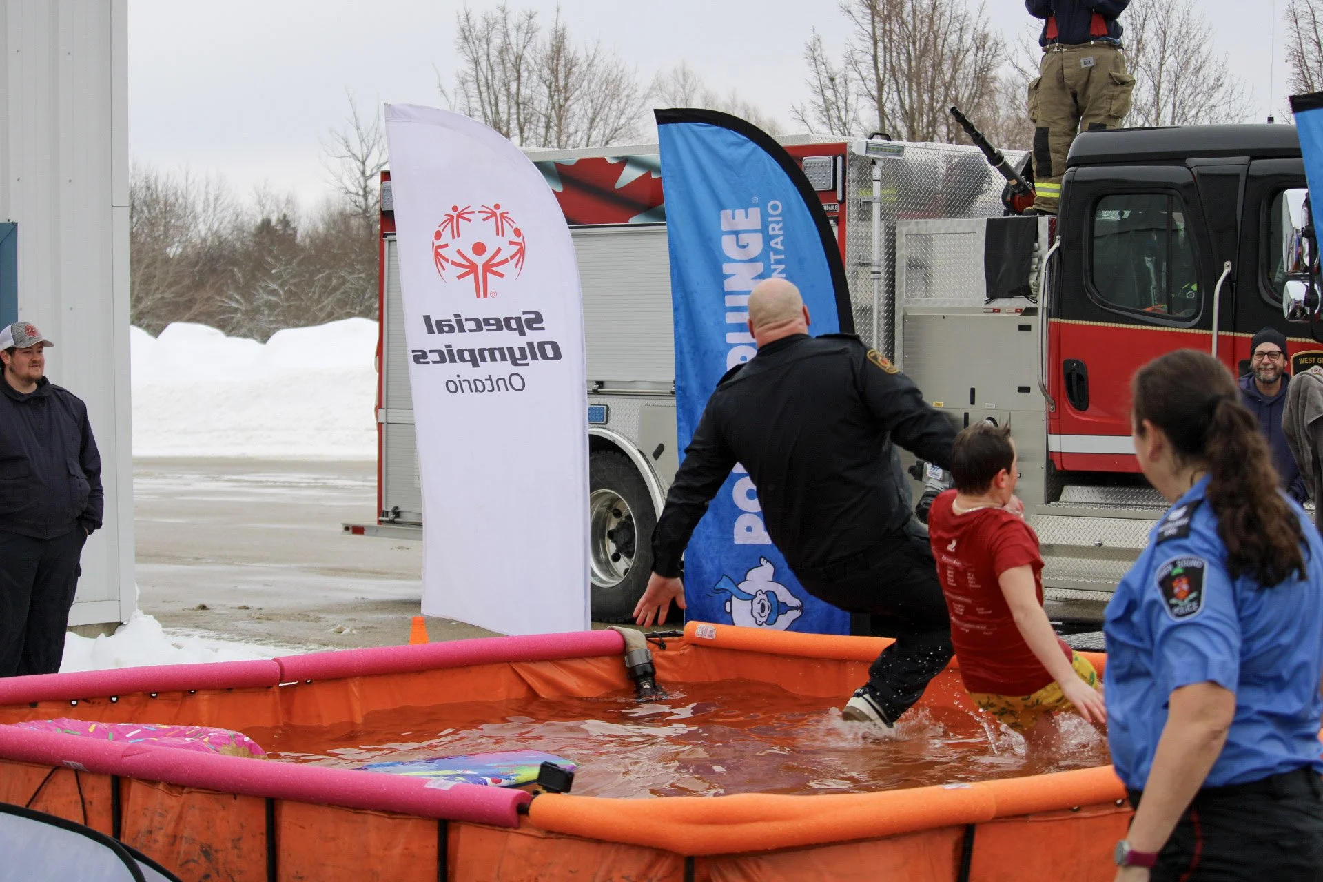 A firefighter in black uniform helping a young boy in red shirt into a pool of water at an outdoor event. Several people, including a woman in a blue uniform and another man in casual clothing, watch nearby. A fire truck with flags stands in the background, with snow on the ground and bare trees.