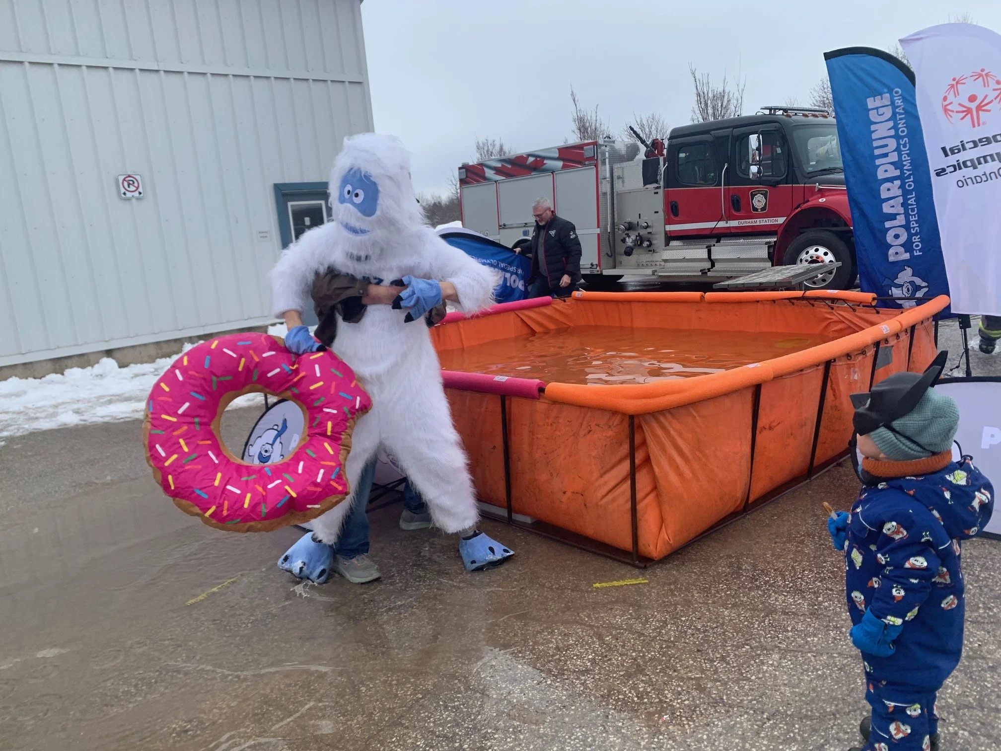 Person dressed in a Yetti costume holding a pink frosted donut floatie with sprinkles, standing next to a small child in winter clothing looking at them, both near an above-ground water tank filled with water, with a fire truck and a blue flag in the background.