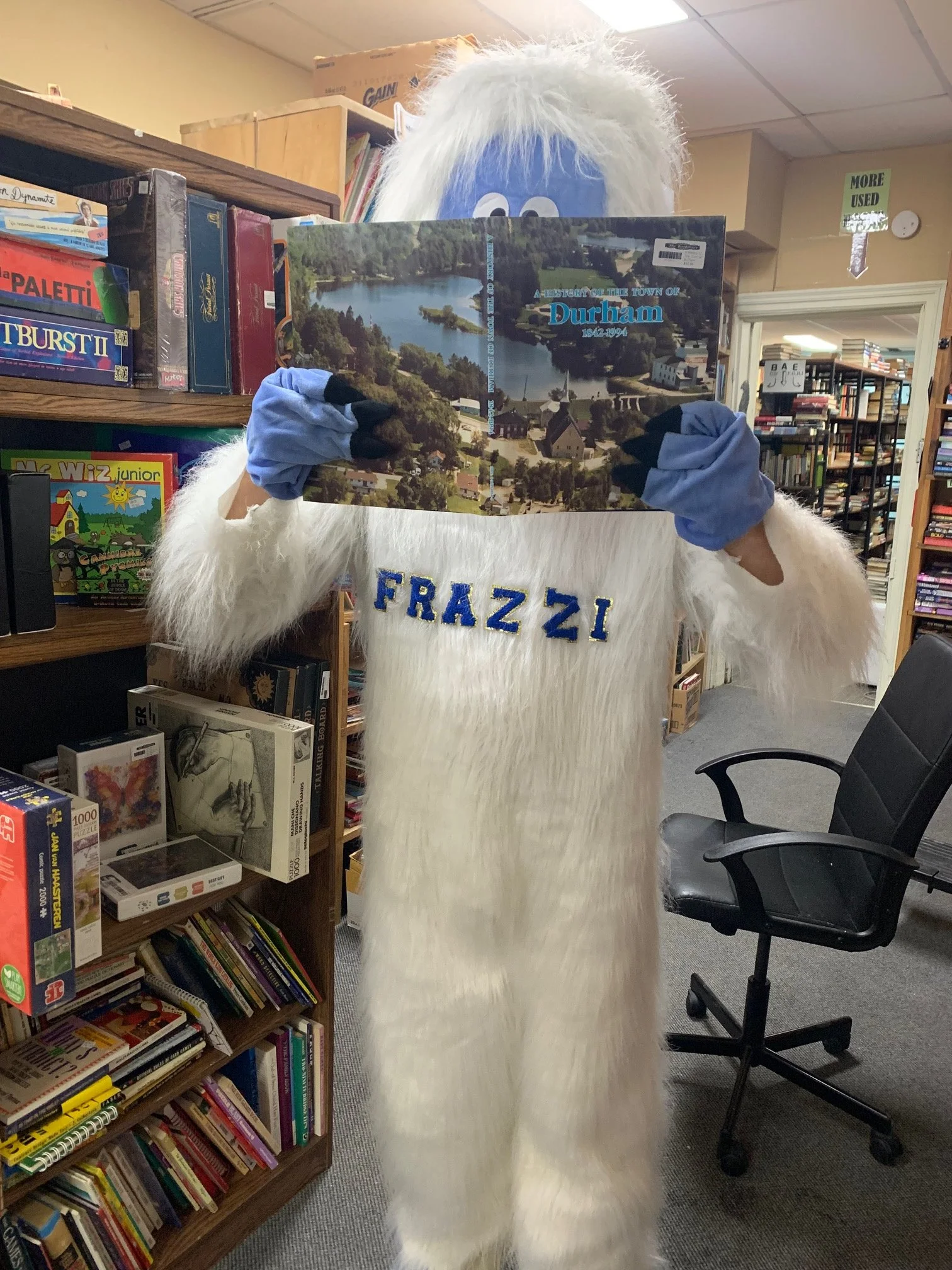 Person dressed in a Bigfoot costume with white fur and blue gloves, holding a book titled "A History of the Town of Durham" in a library.