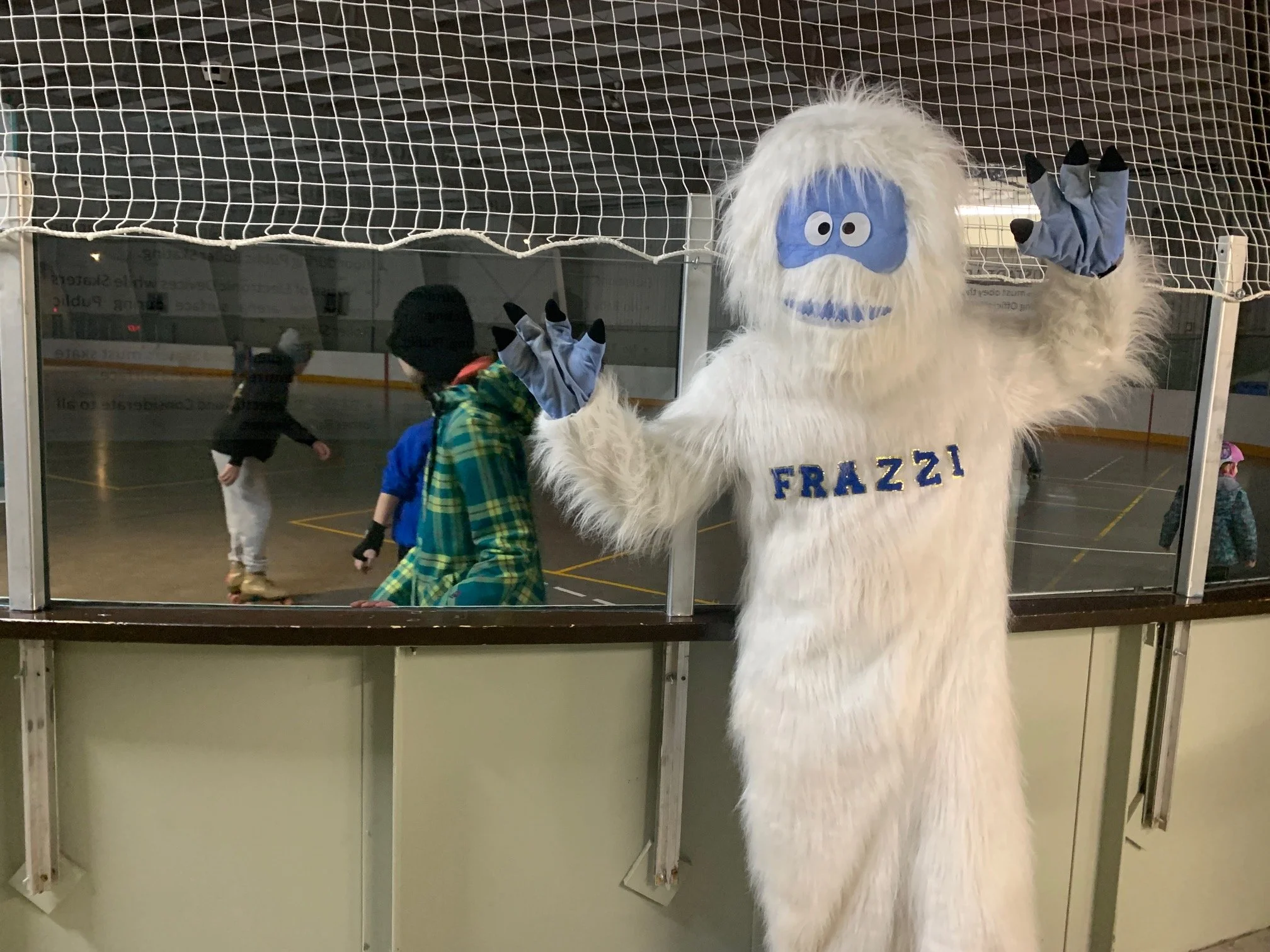 A person dressed in a white yeti costume with blue face paint and the name tag "FRAZZI" stands in front of an ice skating rink, waving with both hands. Children skate in the background.