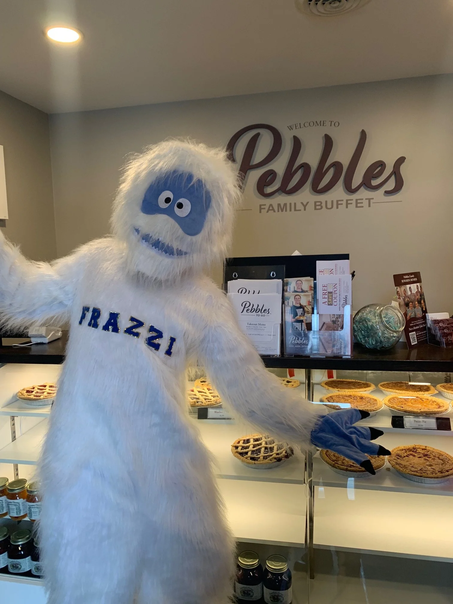 A person in a Bigfoot costume with white fur and a blue face, wearing a shirt that says 'FRAZZI,' standing next to a display of pies at Pebbles Family Buffet.