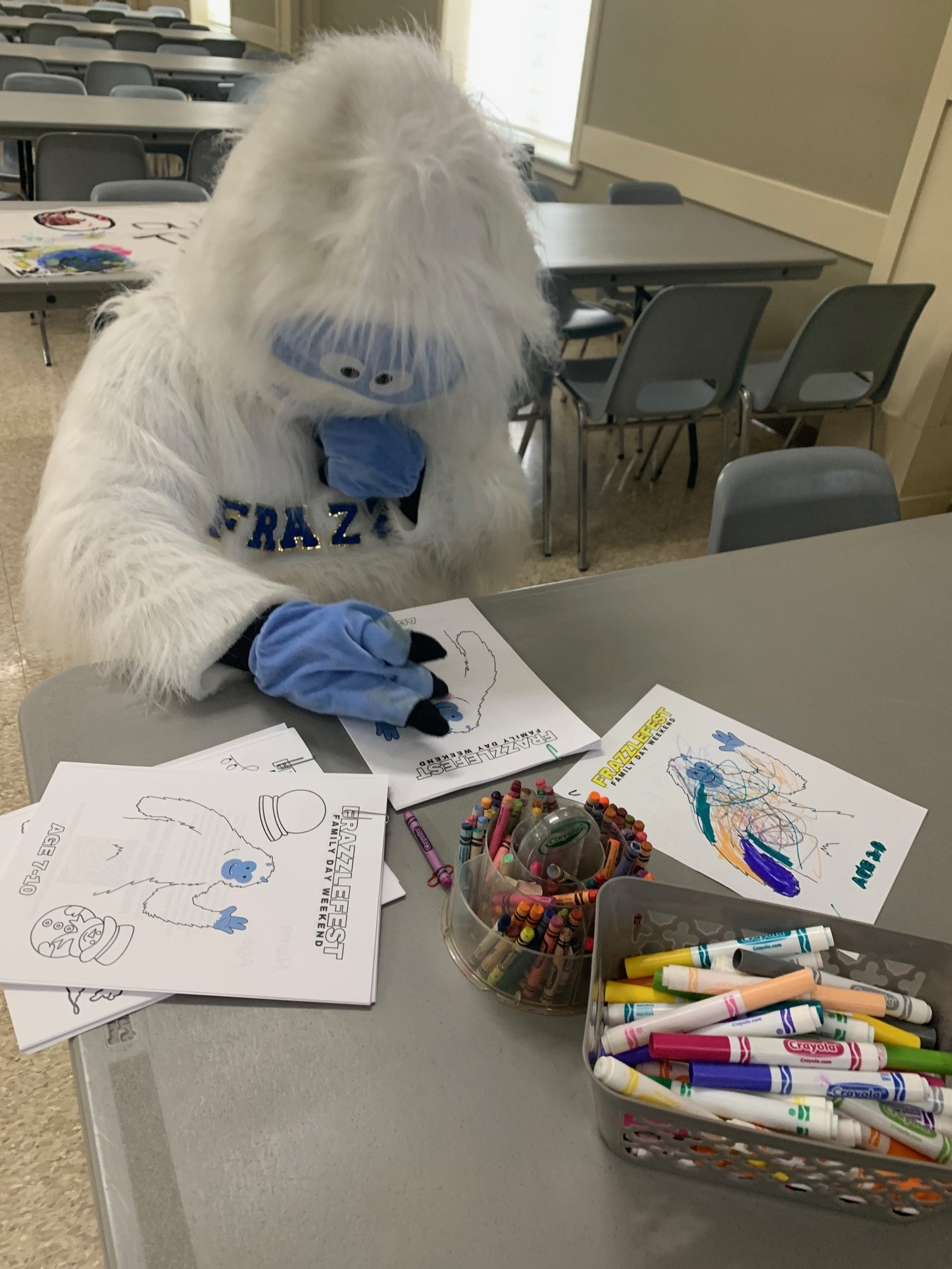 A person dressed in a Sasquatch costume, with a furry white suit and a mask, is coloring a drawing of a Sasquatch on paper. The table has various coloring supplies, including markers and crayons, and other coloring sheets are visible.