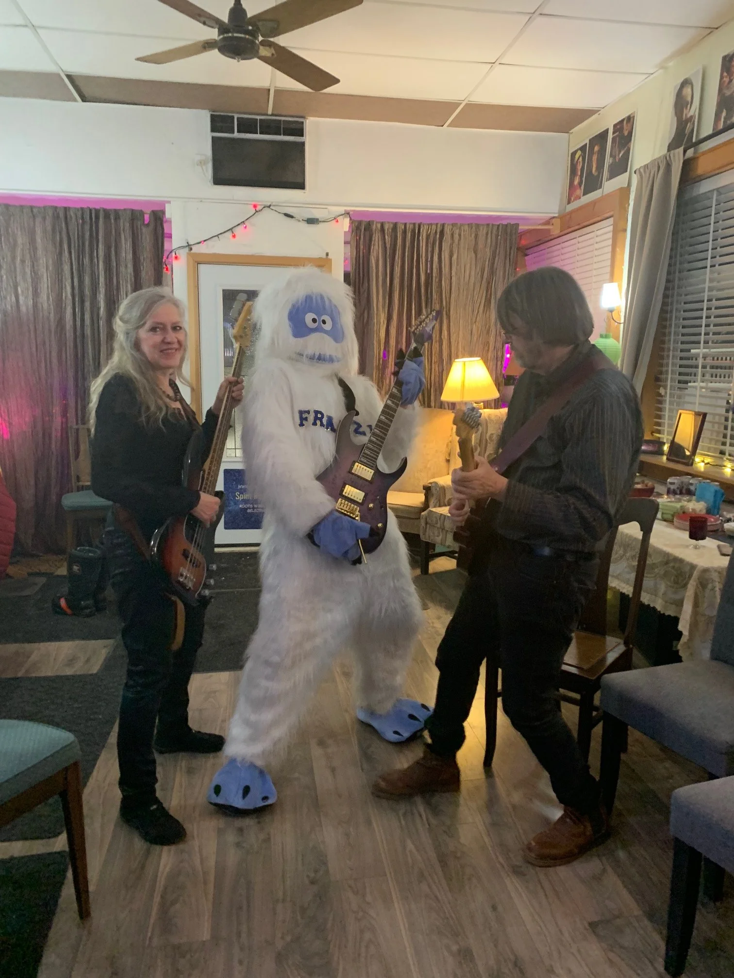Three people holding electric guitars in a room with purple and yellow lighting, decorations, and framed photos on the wall.