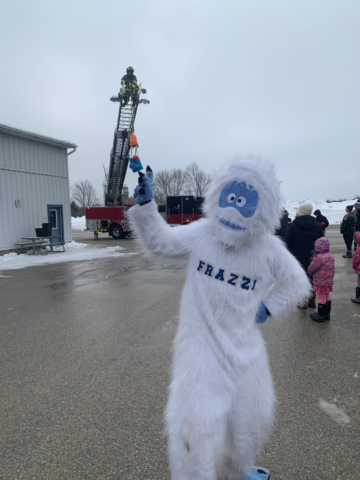Person in a Bigfoot costume with 'FRAZZI' written on it, pointing with one finger, in a snowy outdoor area with fire trucks and a firefighter on an extendable ladder, and several children and adults in the background.