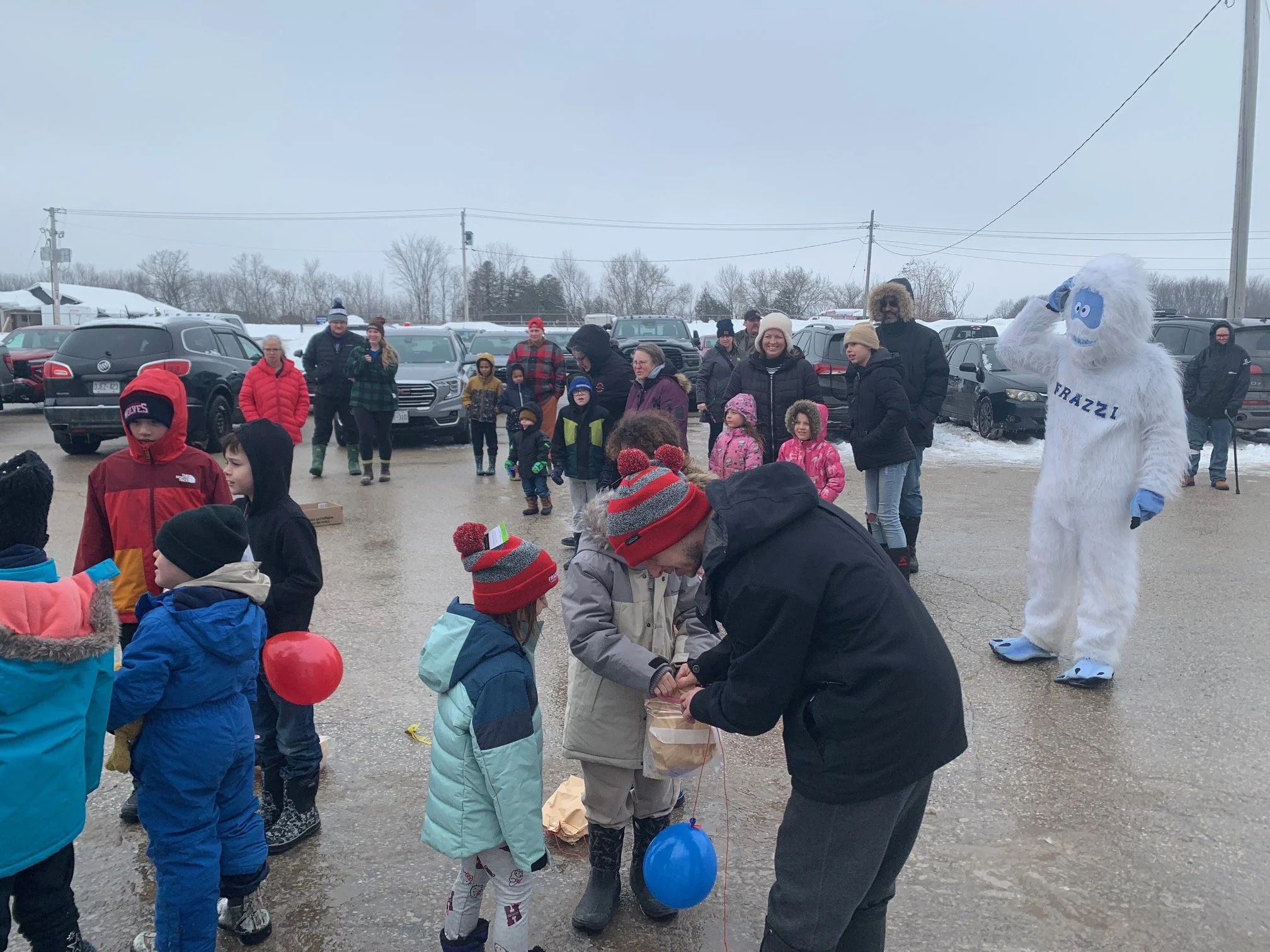 Children and adults gather in a snowy parking lot for a winter event, with a person in a Frazzi mascot costume in the background and a man handing out items to children.