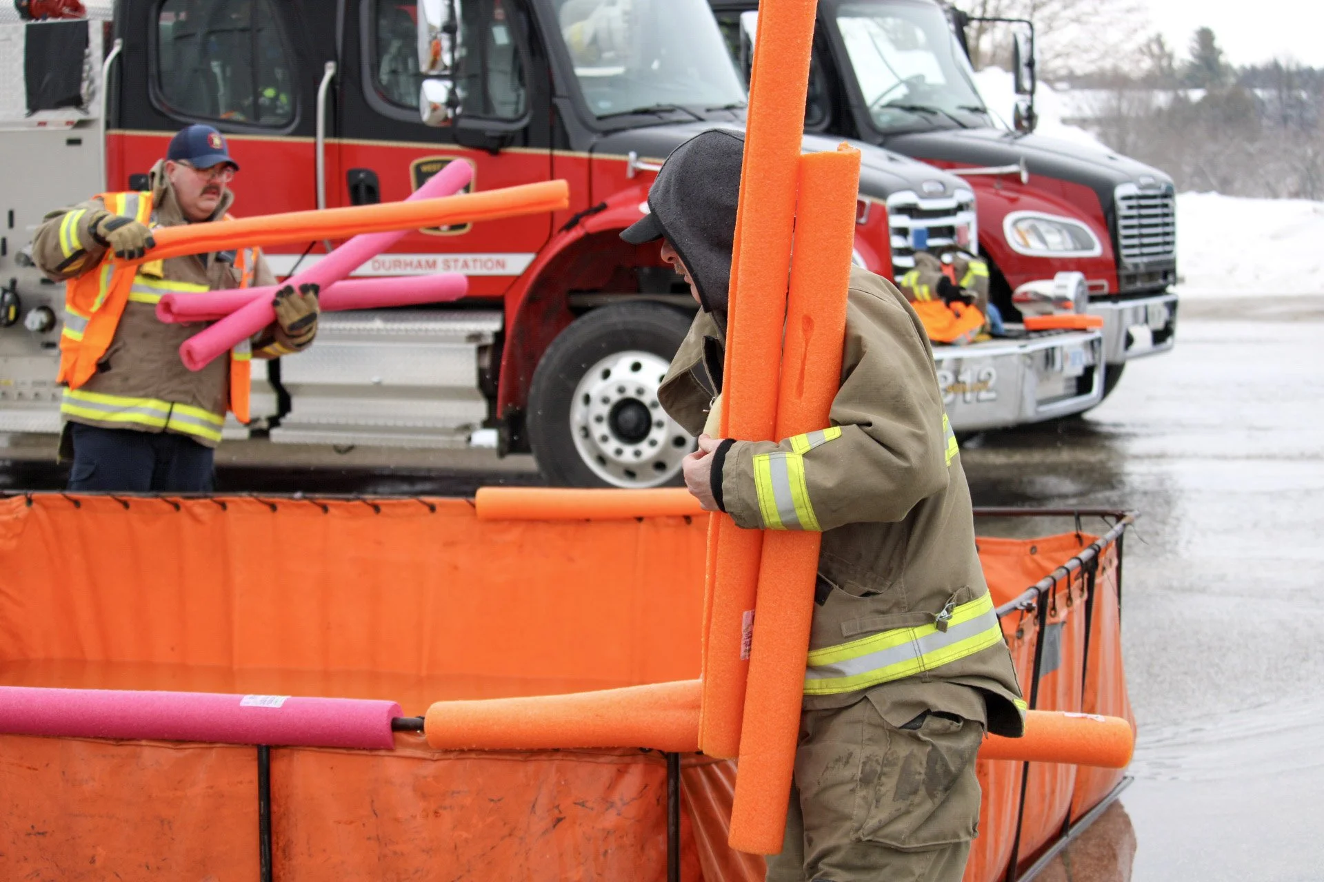 Firefighters are setting up orange and pink flotation devices around a water rescue training pool, with fire trucks in the background on a rainy day.