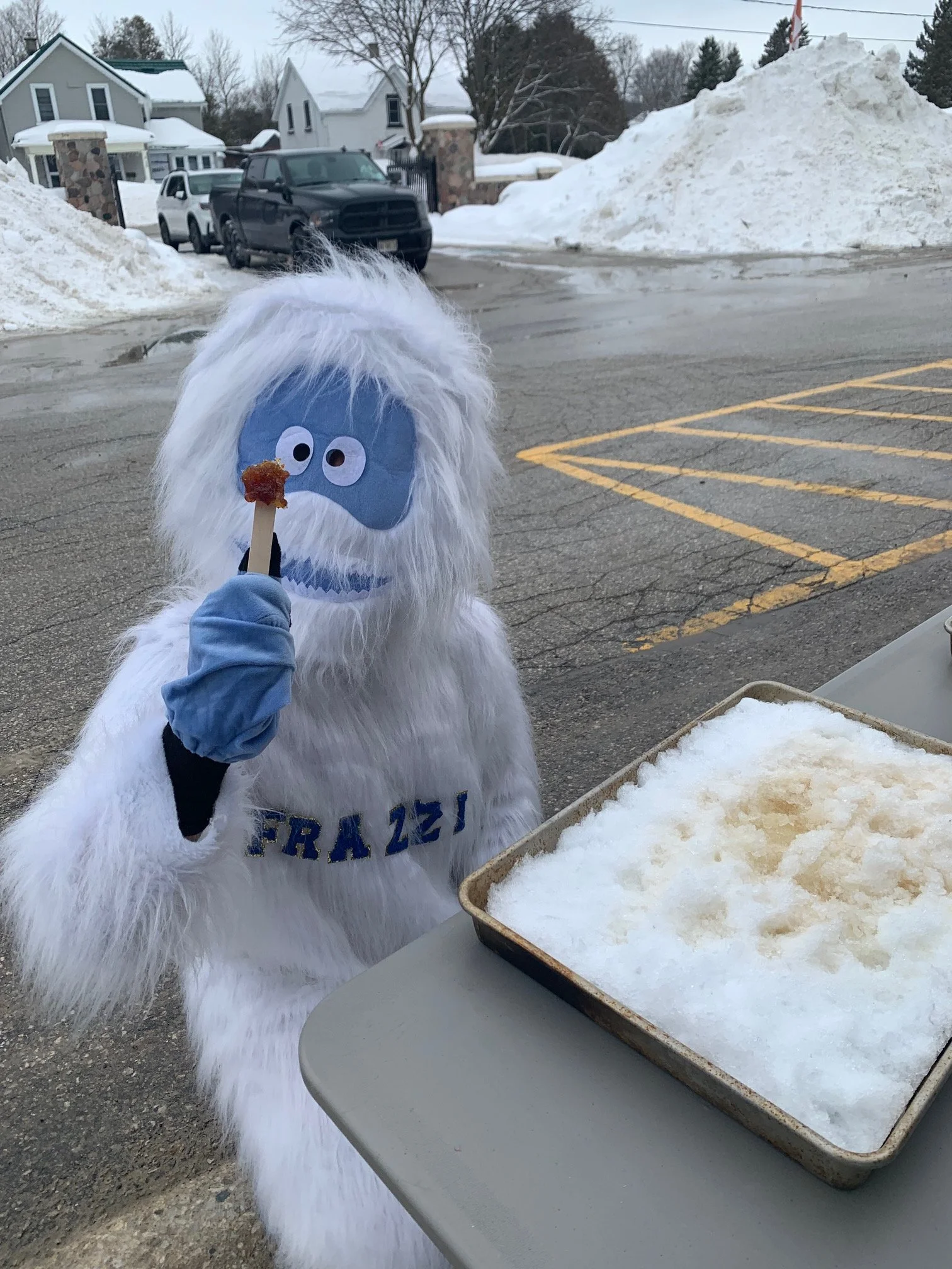 Person dressed in a Yeti costume holding a wooden stick with a piece of meat, standing next to a tray of snow.