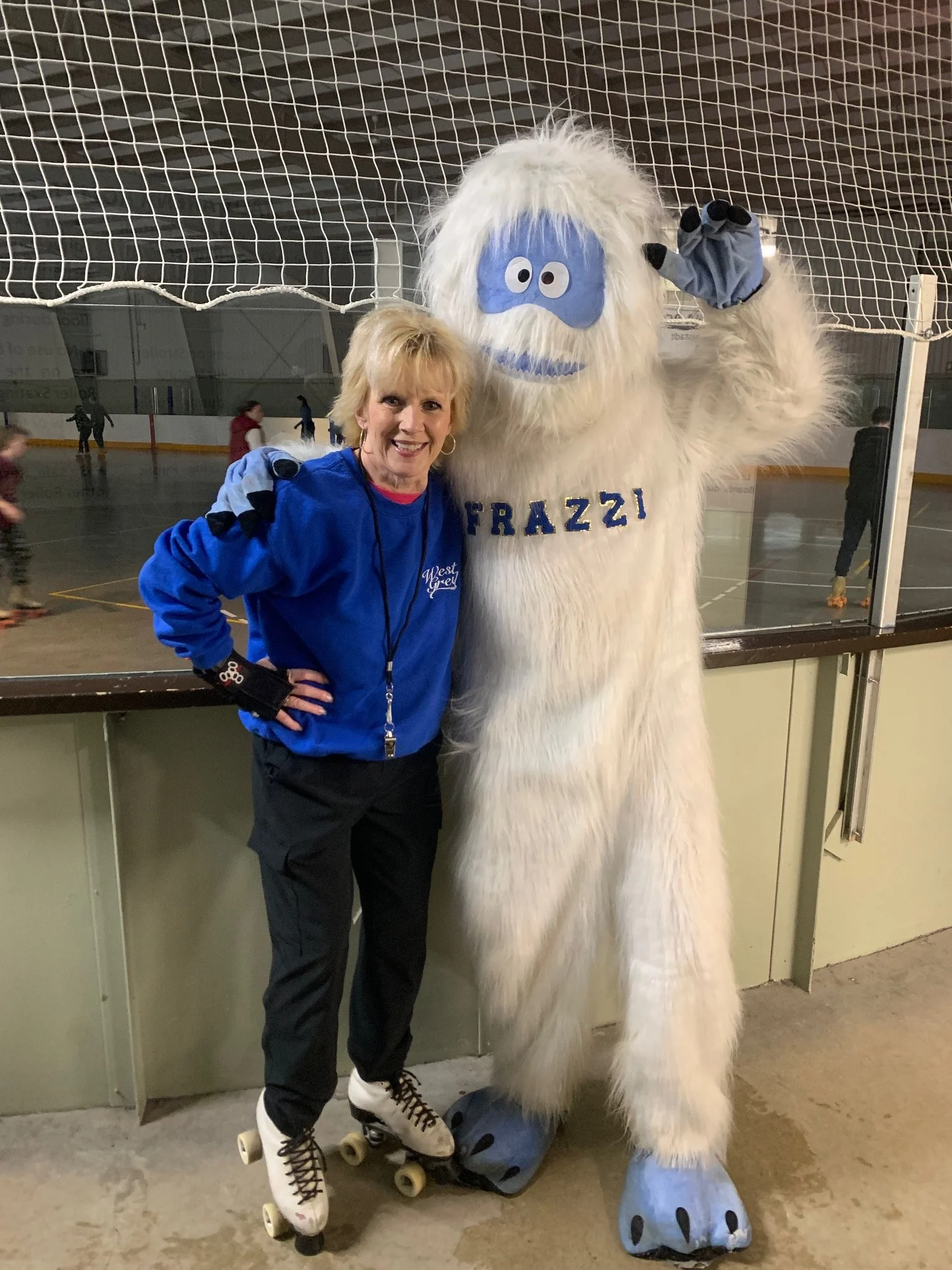 A woman in a blue sports jacket and roller skates standing next to a large, furry, white monster mascot with blue face paint and the word 'FRAZZI' on its chest, in an indoor ice skating rink.