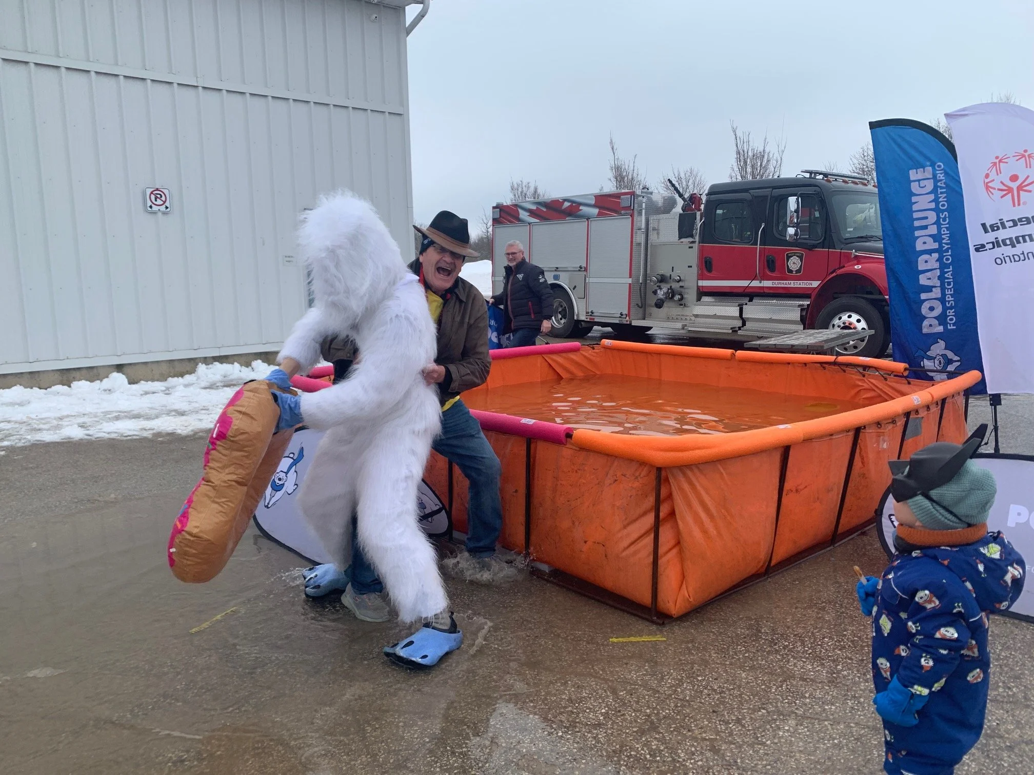 A person in a suit and fur costume playfully splashes into a water-filled orange pool with a firefighter's hat on, while another small child watches nearby. A fire truck and blue promotional flags are in the background, and snow is on the ground.