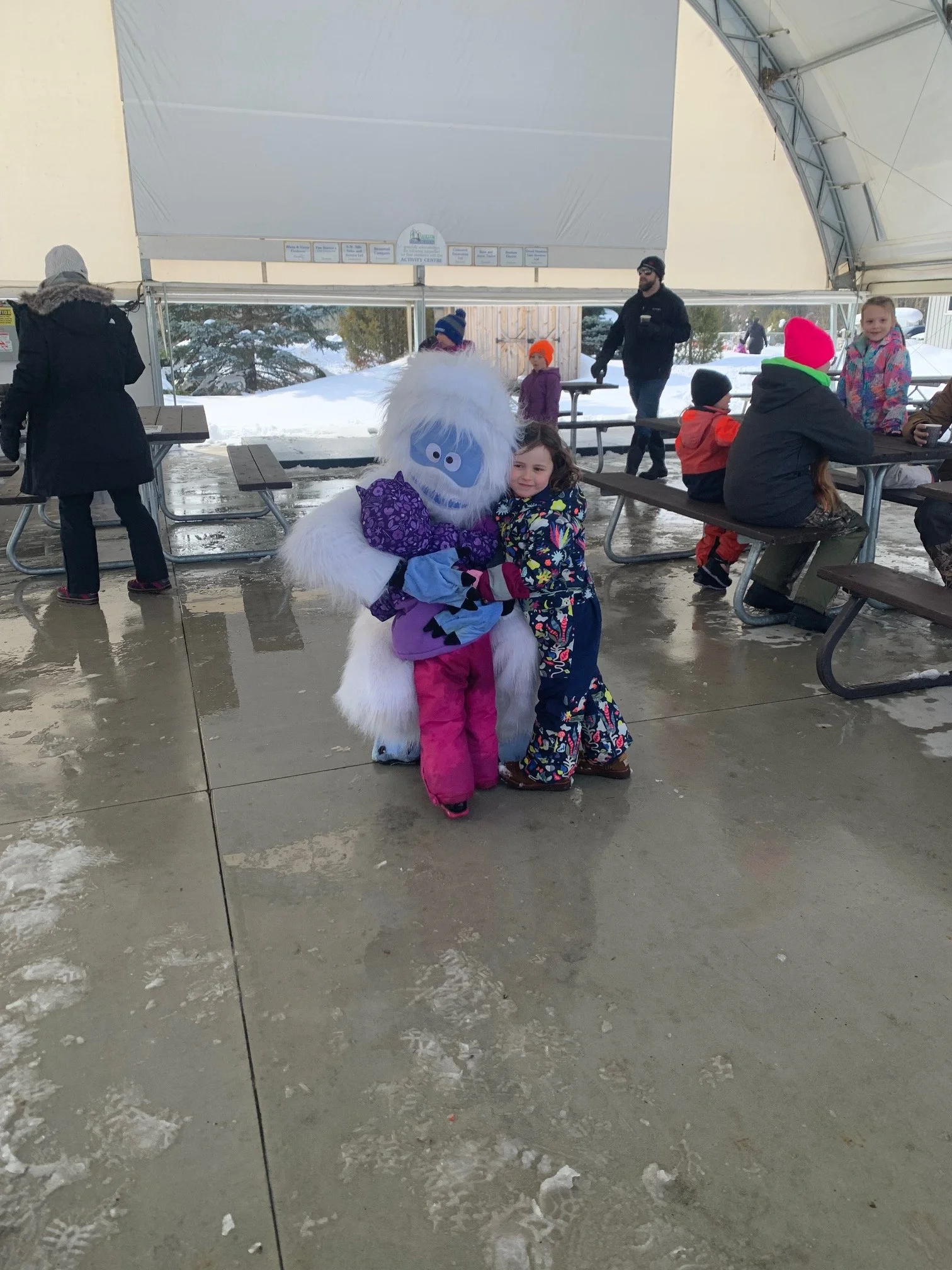 Two children in colorful winter clothing hugging a person in a large, furry, monster-like costume inside a covered area with snow outside.