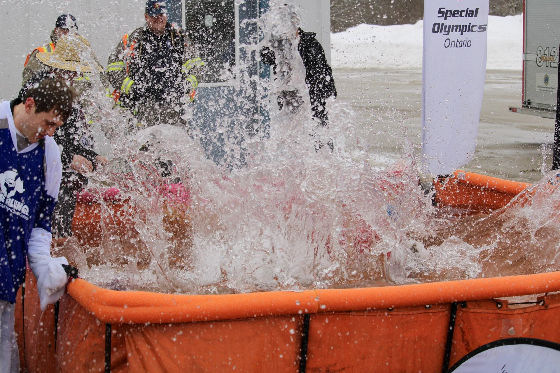 People participating in a special Olympics event on an outdoor ice or snow surface. Some are in firefighter gear, and others are near an orange water or ice rescue pool, with water splashing out. A sign reads "Special Olympics Ontario."